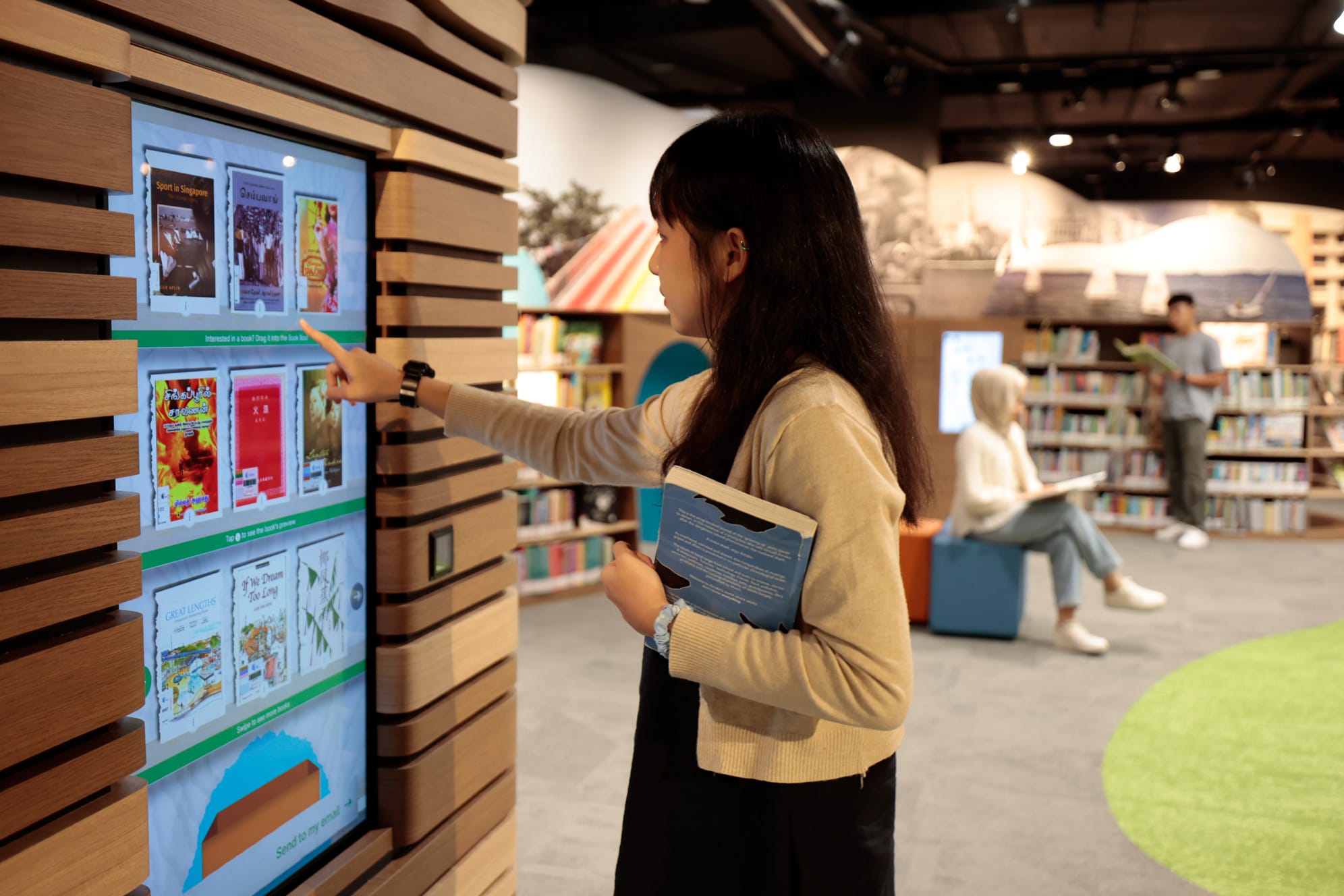 A person touches a large interactive display in a modern library, while holding a book; other patrons are reading in the background.