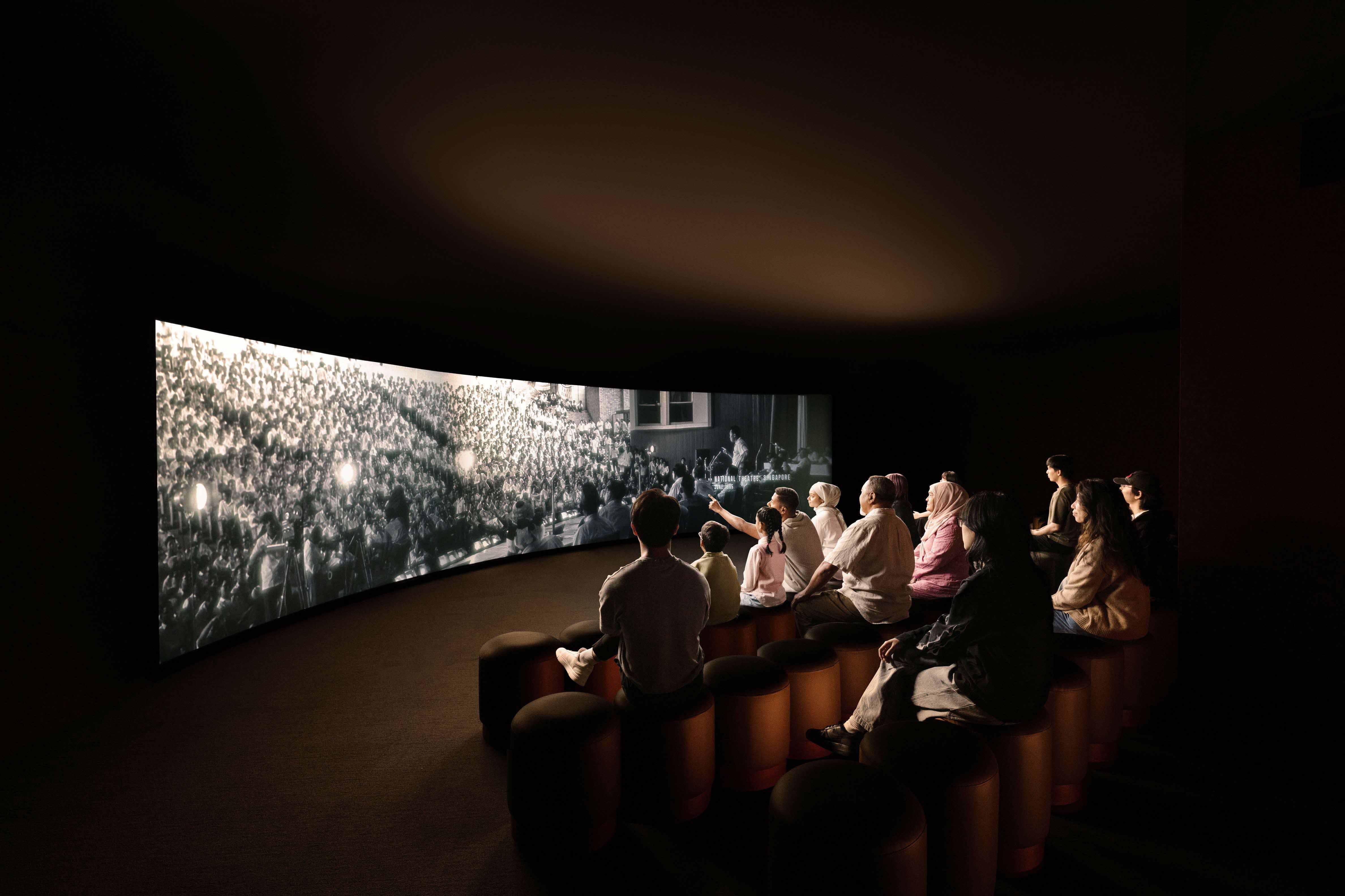 A group of people sit on stools in a dark room, watching a large black-and-white screen showing a crowded event.