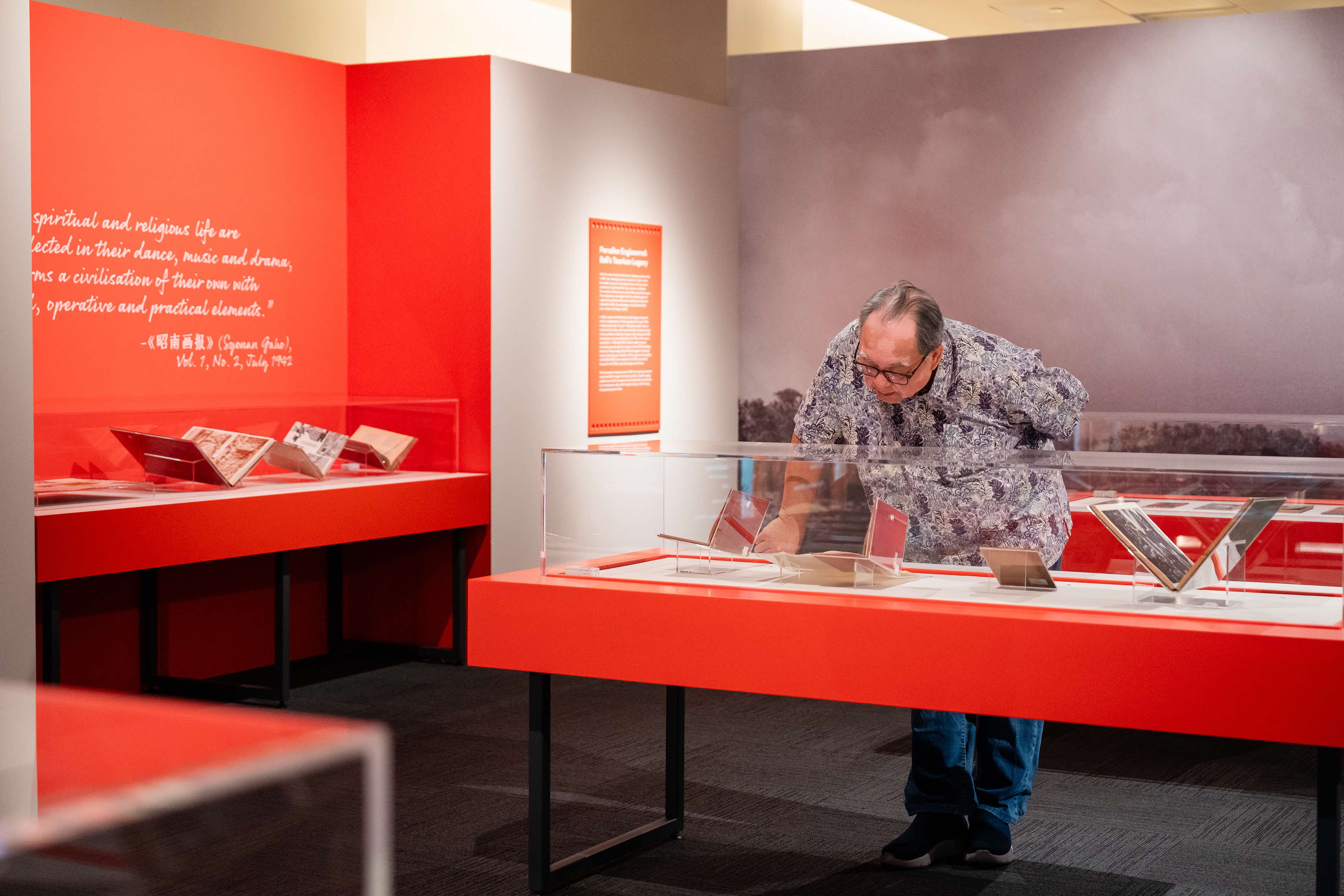 A person is examining items in a museum display case with red exhibit tables and informational text on the walls.