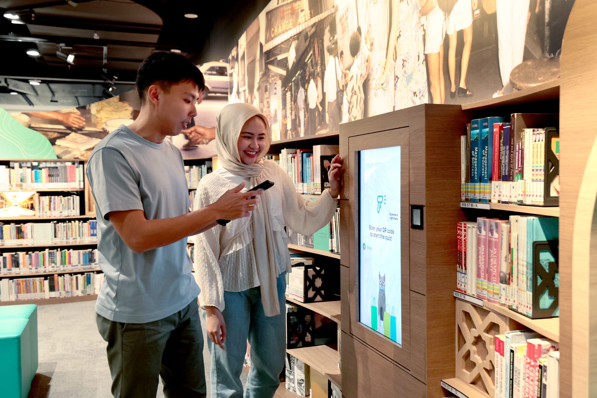 Two people interact with a touch screen in a library, surrounded by shelves of books and wall murals.