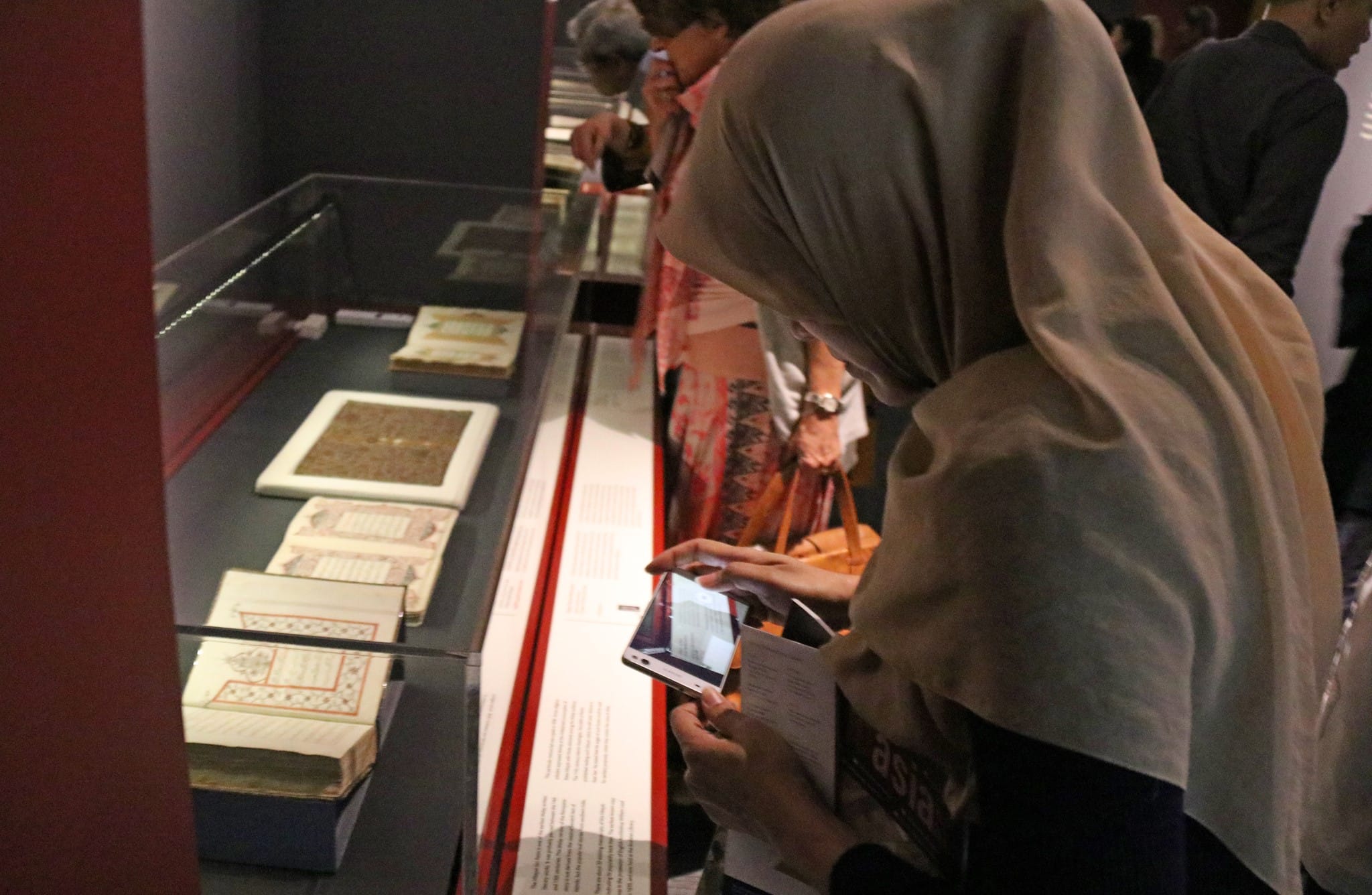 A person wearing a hijab is photographing ancient manuscripts displayed in a glass exhibit case.