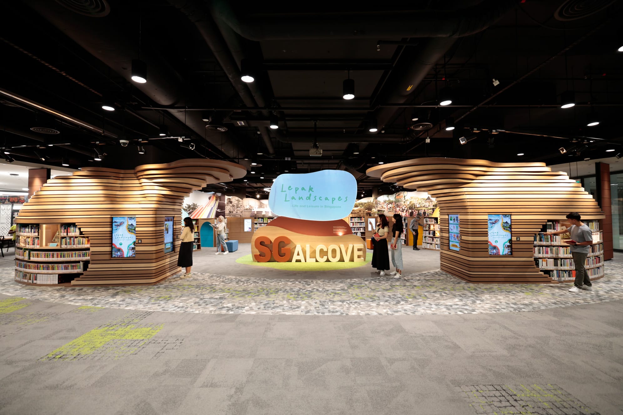 A modern library display with curved wooden shelves, digital screens, and a large sign reading "SG Alcove."