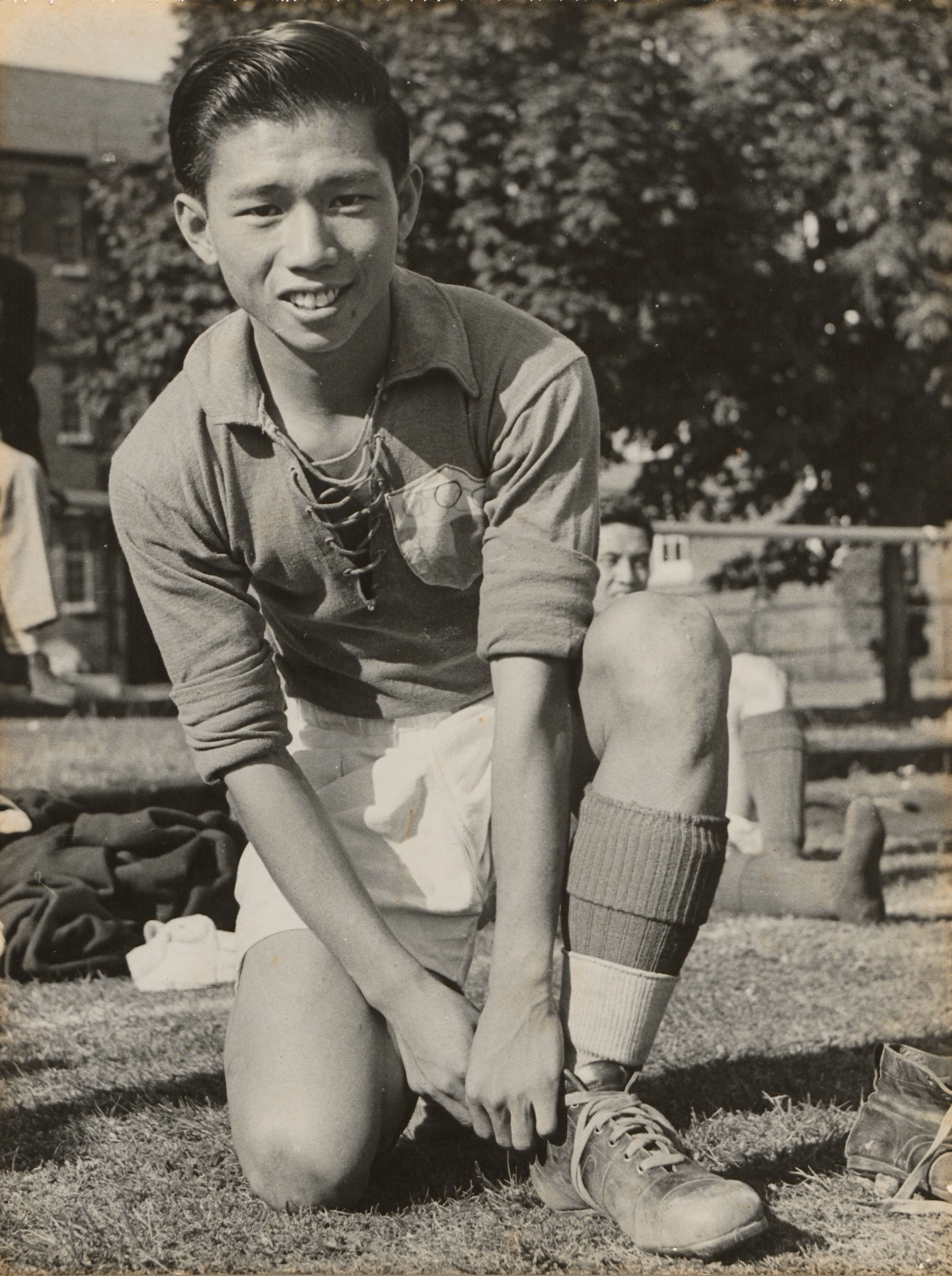 A soccer player kneels on the grass, tying his shoe while wearing a lace-up jersey, shorts, and socks.