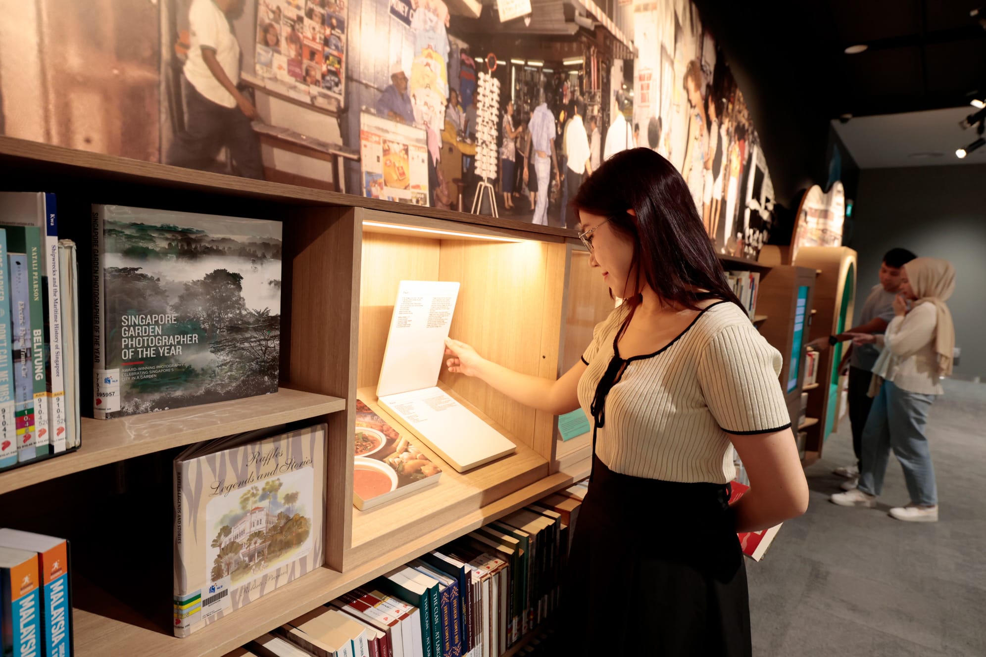 A person is reading a book in a library with wooden shelves and various books on display.