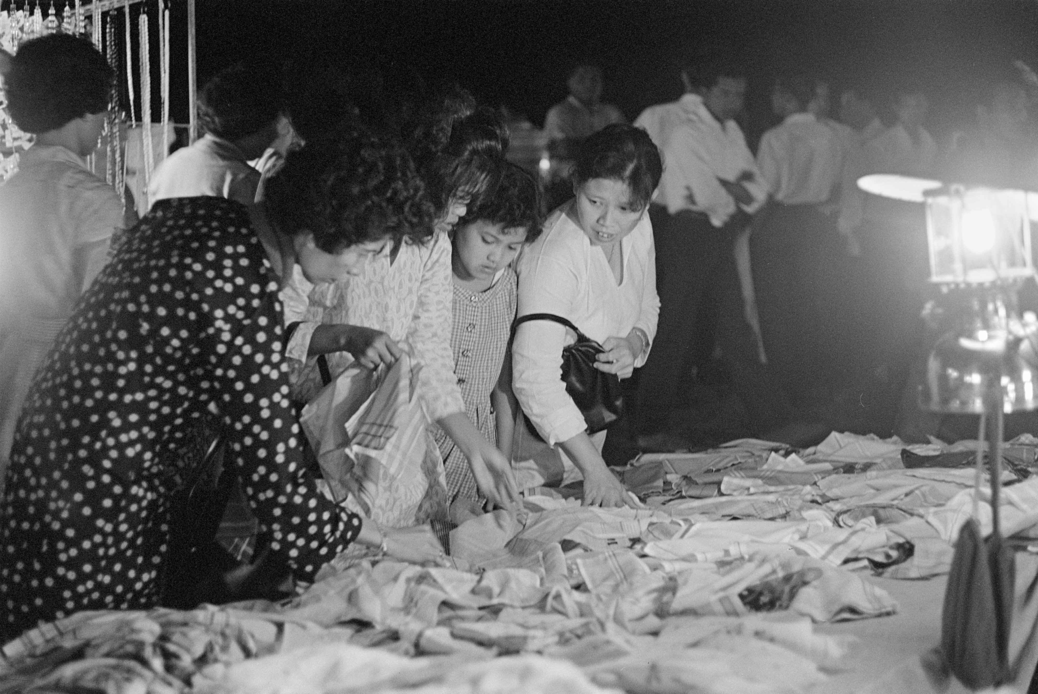 People sorting through clothes on a table at an outdoor nighttime event with a bright lamp illuminating the area.