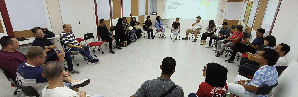 A group of people sits in a circle in a brightly lit room with "Learning Space" visible on the wall.