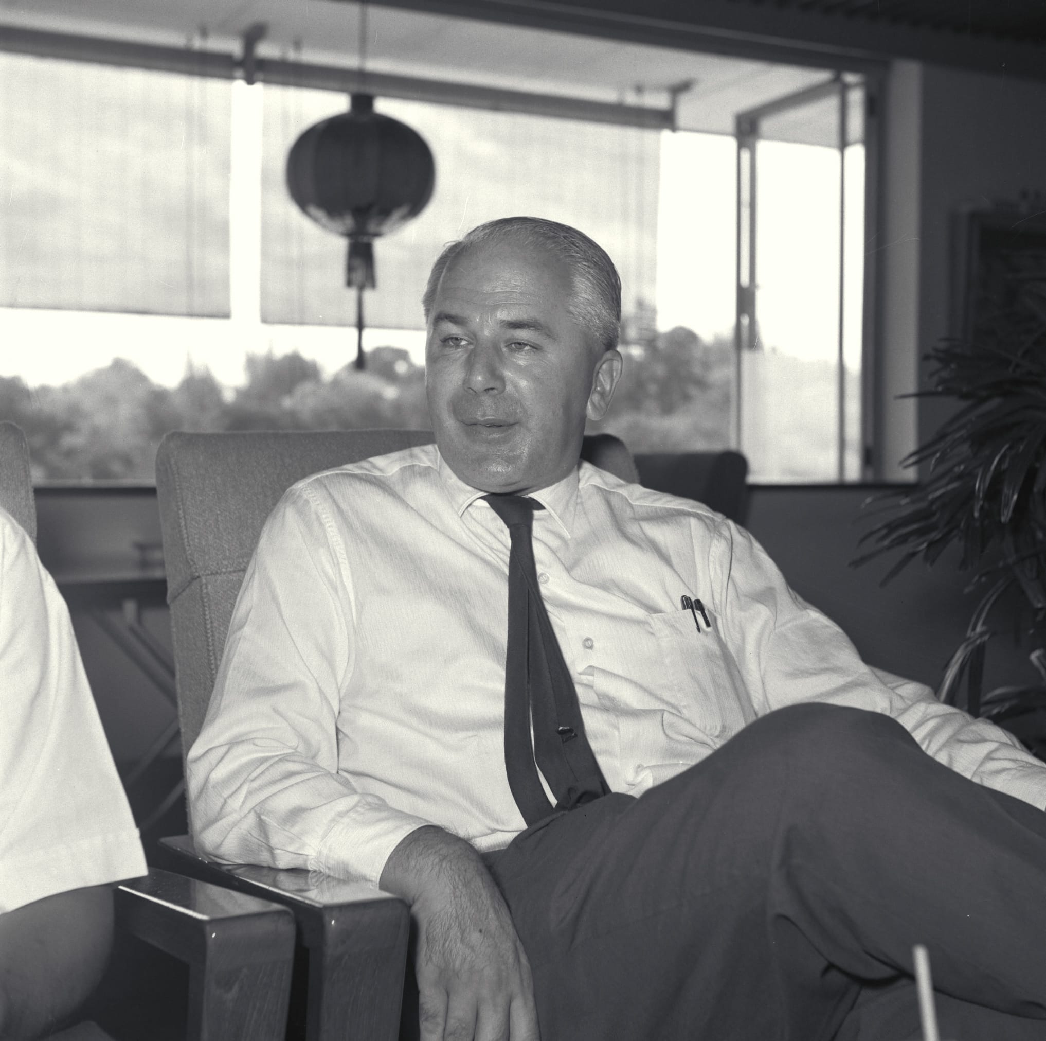 A person wearing a dress shirt, tie, and pen in pocket is seated indoors with an open window and a lantern in the background.