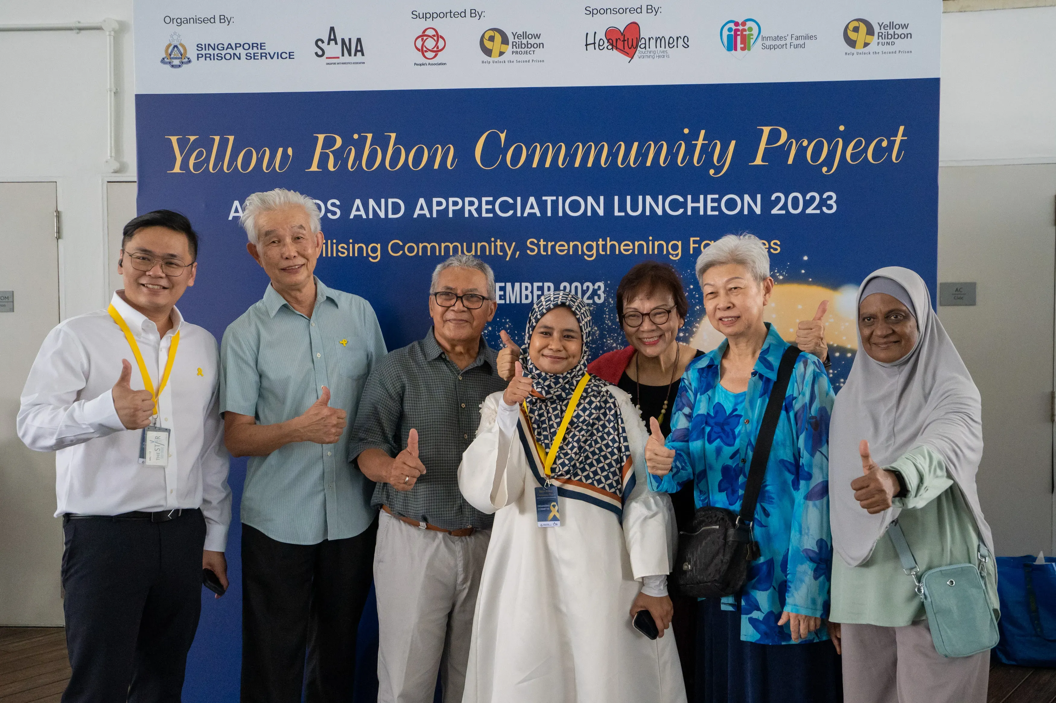 Seven people pose smiling with thumbs up before a Yellow Ribbon Community Project luncheon 2023 banner