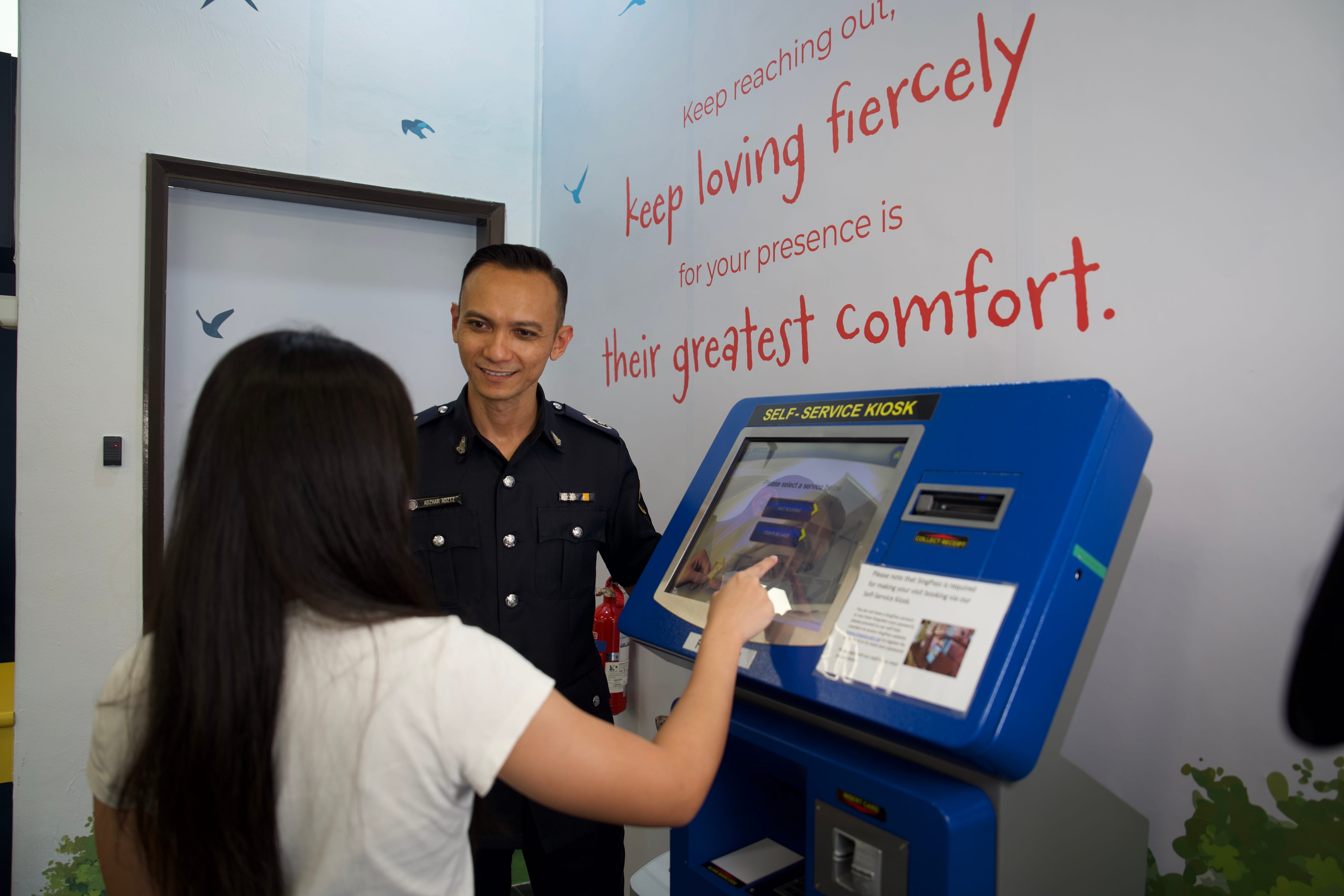 A woman in a white top touches the screen of a blue self-service kiosk while a male prison officer stands beside her, smiling. Behind them, a wall decorated with bird silhouettes reads: “Keep reaching out, keep loving fiercely for your presence is their greatest comfort.”