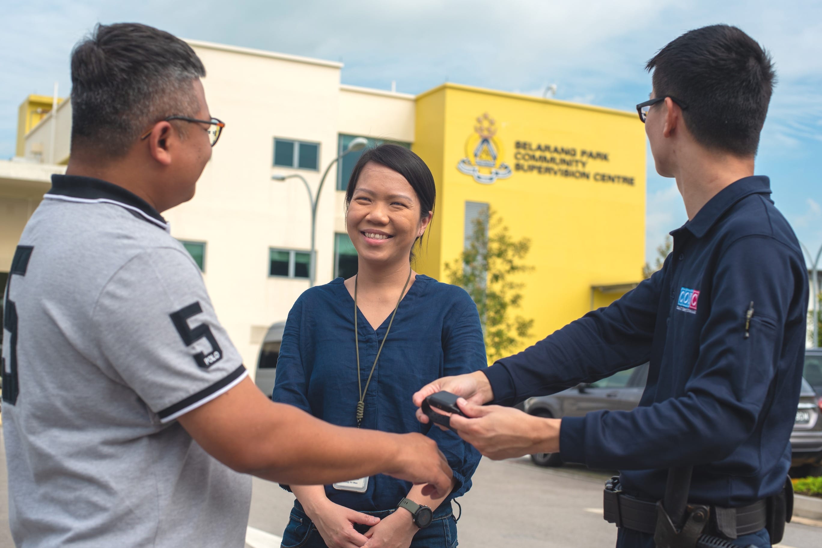 Three people talk outside a building; a uniformed man hands a device to a man as a woman smiles.