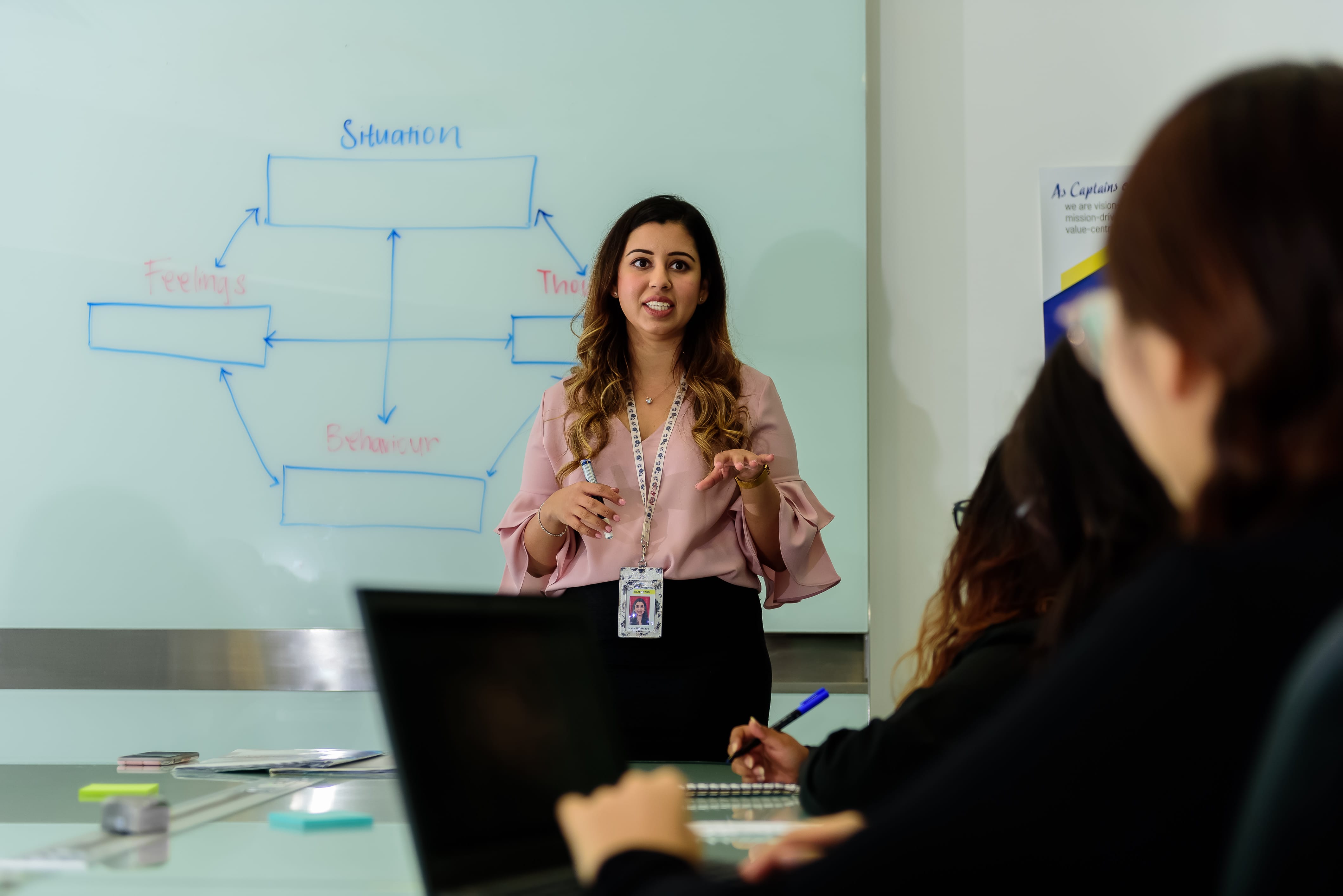 A psychologist in a pink blouse stands at the front of a meeting room, speaking and gesturing to a seated group. Behind her, a whiteboard displays a diagram linking “Situation”, “Feelings”, “Thoughts” and “Behaviour”. Participants in the foreground take notes beside an open laptop.