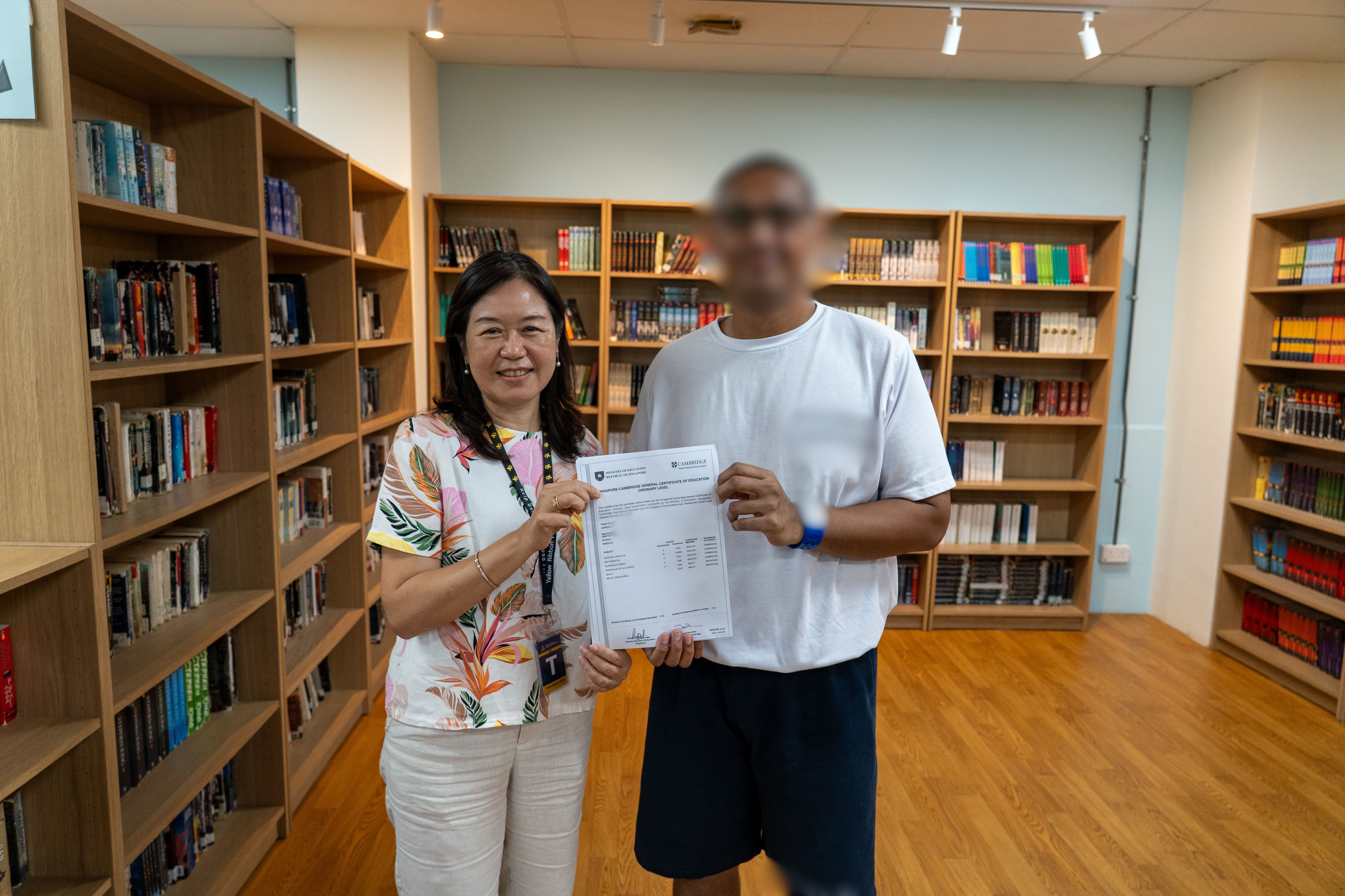 A smiling woman in a floral blouse stands beside an inmate, whose face is blurred. They jointly hold up a Singapore-Cambridge GCE Ordinary Level certificate towards the camera. Wooden shelves filled with books surround them in a brightly lit library.
