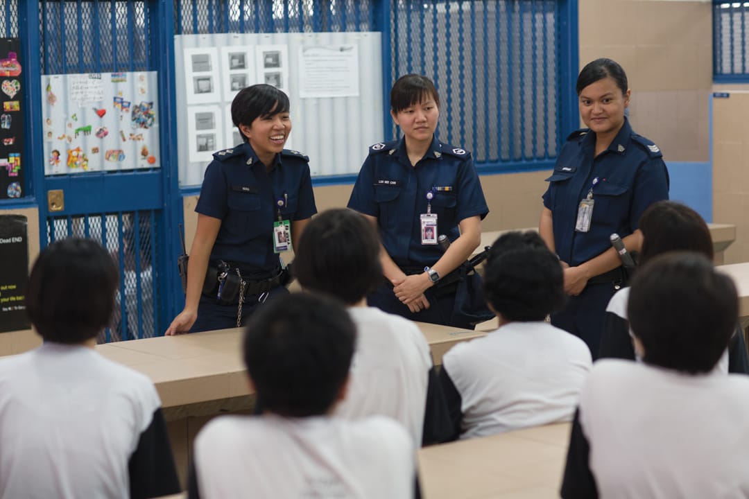 Three uniformed officers stand before a group of seated individuals, with bulletin boards and blue metal screens in the background.