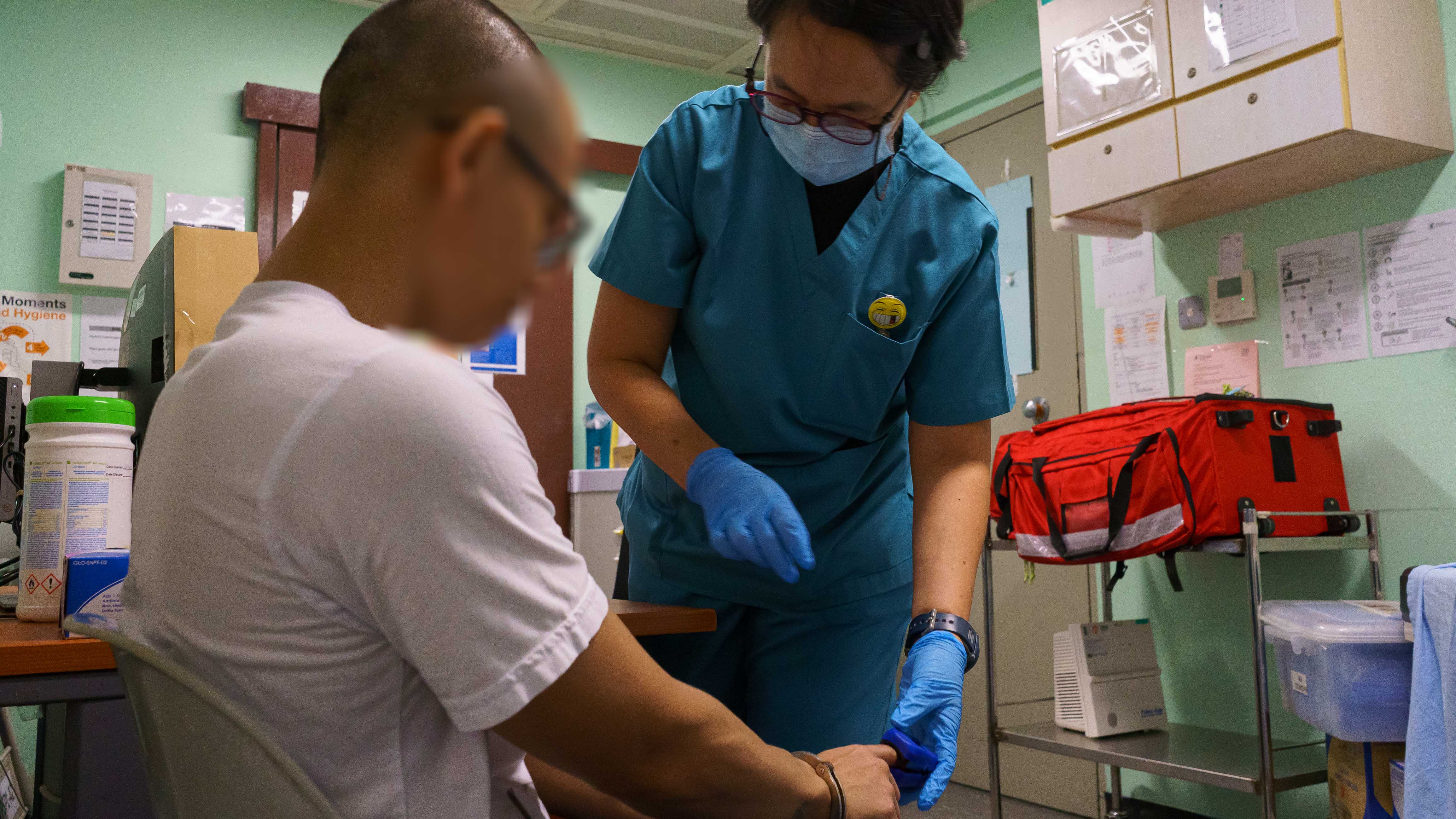 A person in blue medical scrubs attends to another person in a room with medical equipment and supplies.