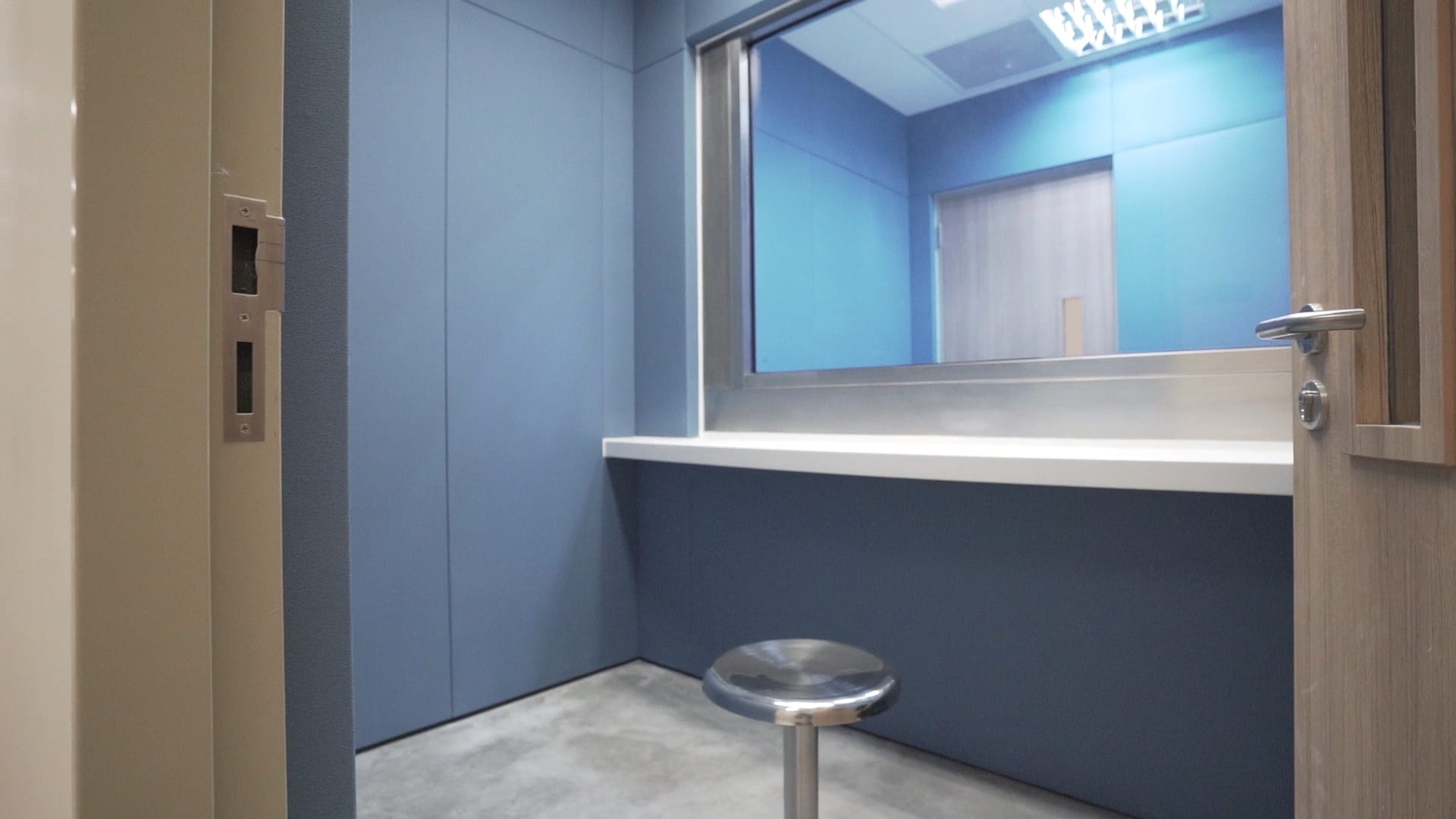 A small blue room with a counter under a large window and a metal stool in the center.