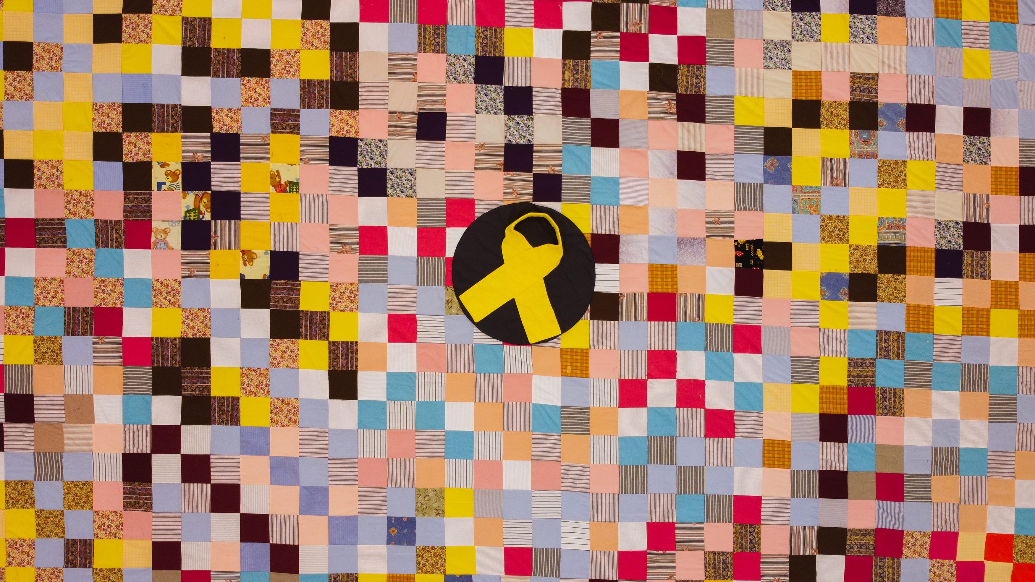 A large patchwork quilt made up of hundreds of colourful fabric squares is displayed flat, creating a vibrant mosaic of different patterns, textures, and colours. At the centre of the quilt is a large yellow awareness ribbon placed on a black circular background, drawing attention as the focal point of the artwork. The quilt includes a mix of floral prints, stripes, solid colours, and patterned fabrics, with some patches featuring children's illustrations. The yellow ribbon symbolises hope, remembrance, and support, while the handmade quilt represents unity, compassion, and the coming together of many individual contributions into a shared message of care and solidarity.