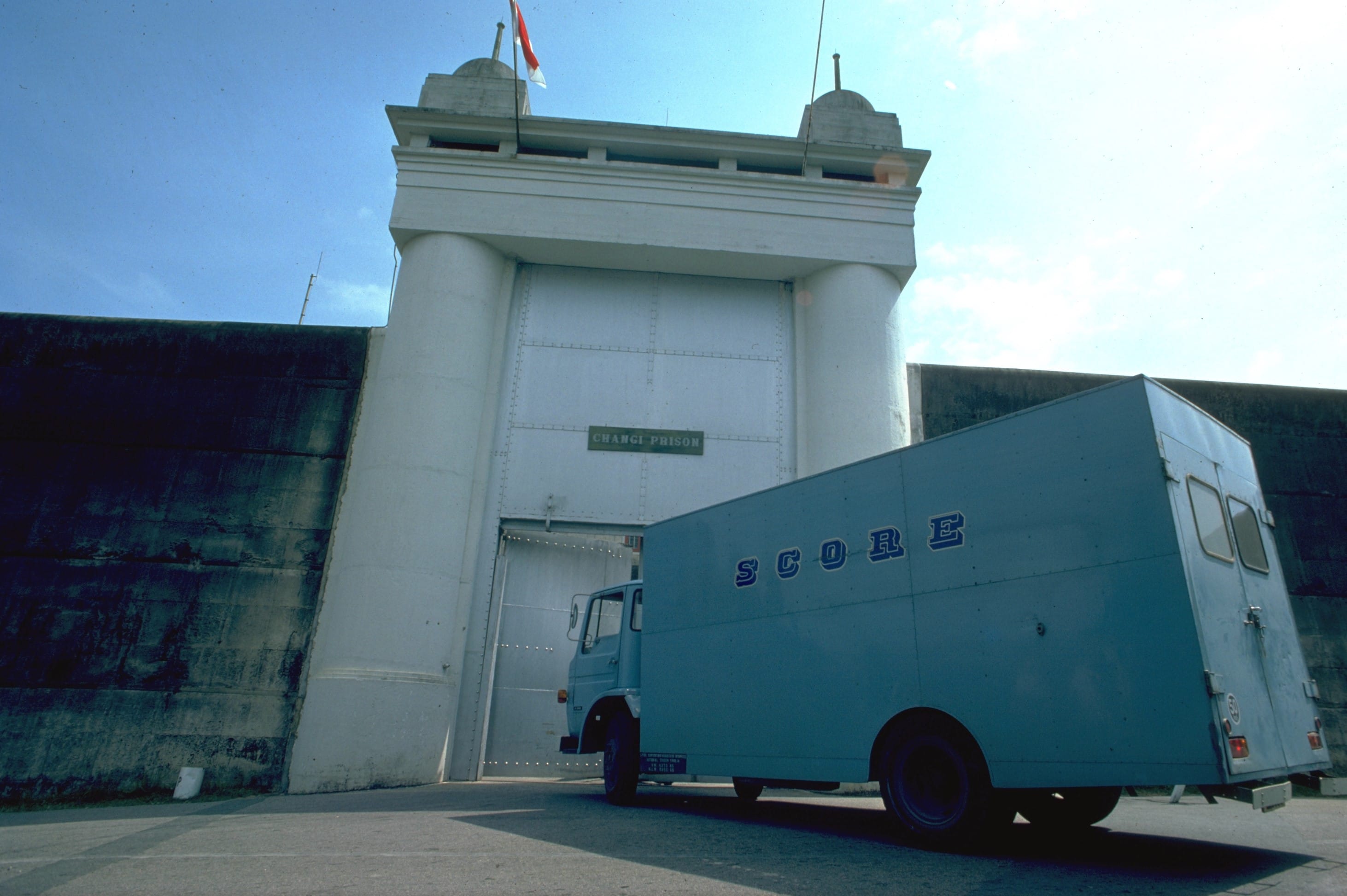 A blue truck with "SCORE" on it is parked in front of a large entrance labeled "Changi Prison."