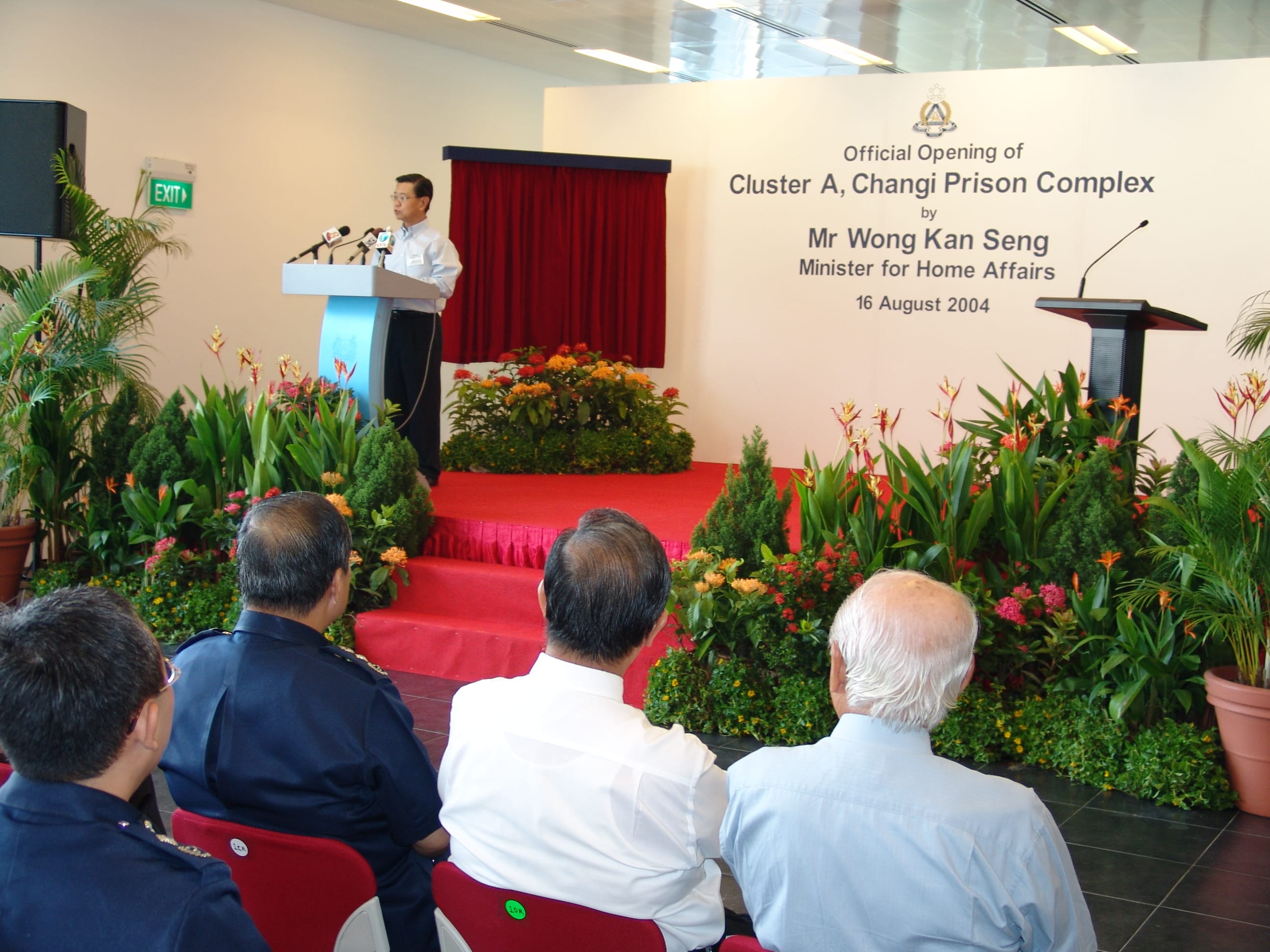 A speaker stands at the podium during an official opening event surrounded by plants; audience in foreground watches.