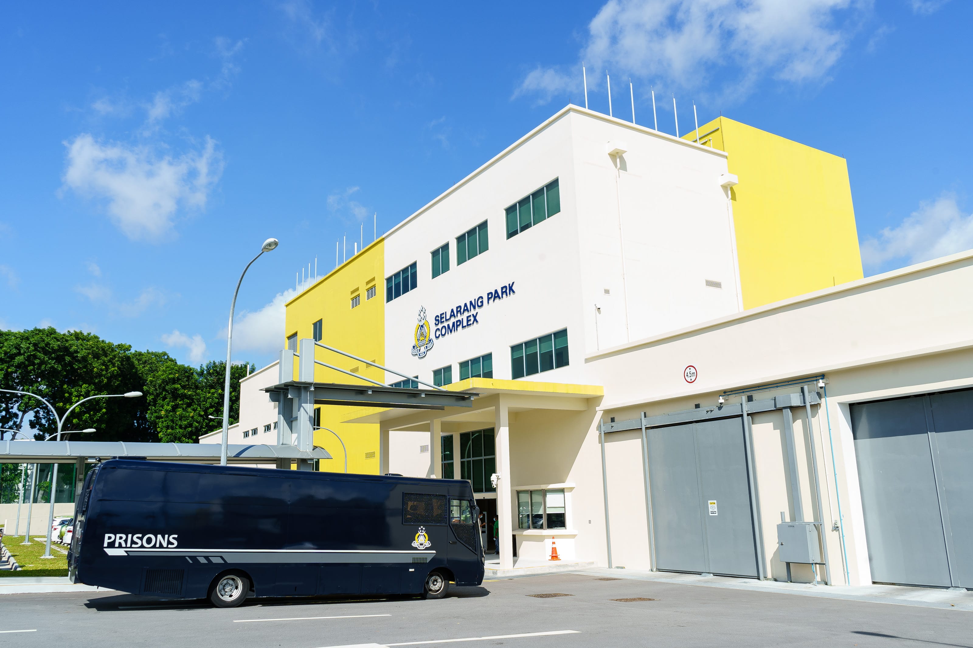 A bright daytime photograph shows the exterior of the Selarang Park Complex, a modern Singapore Prison Service facility with white and yellow buildings set against a vivid blue sky with scattered clouds. The building's name and Singapore Prison Service emblem are prominently displayed on the façade. In the foreground, a dark blue prison transport vehicle marked "PRISONS" is parked outside the entrance, while large secure gates and perimeter access points are visible on the right side of the building. The clean, well-maintained surroundings, along with the secure infrastructure, convey the facility's role as a correctional institution.