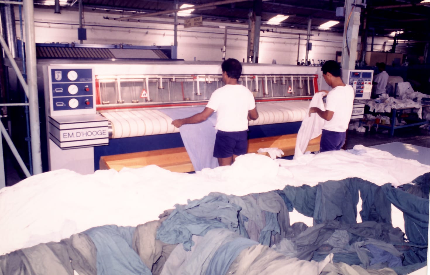 Two men operate a large ironing machine in a laundry facility, surrounded by piles of clothes.