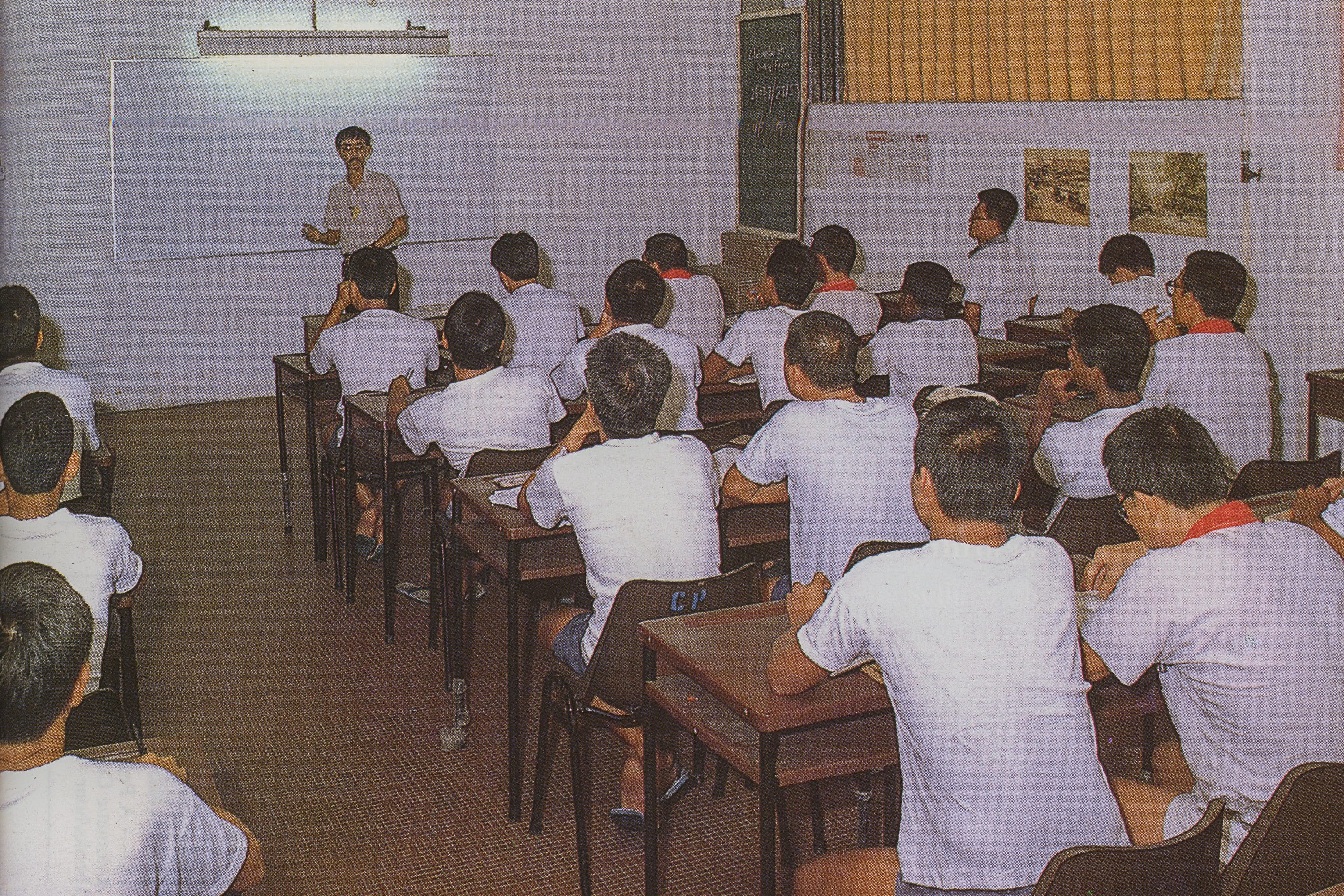 A classroom with students wearing white shirts sitting at desks, facing a teacher standing near a whiteboard.