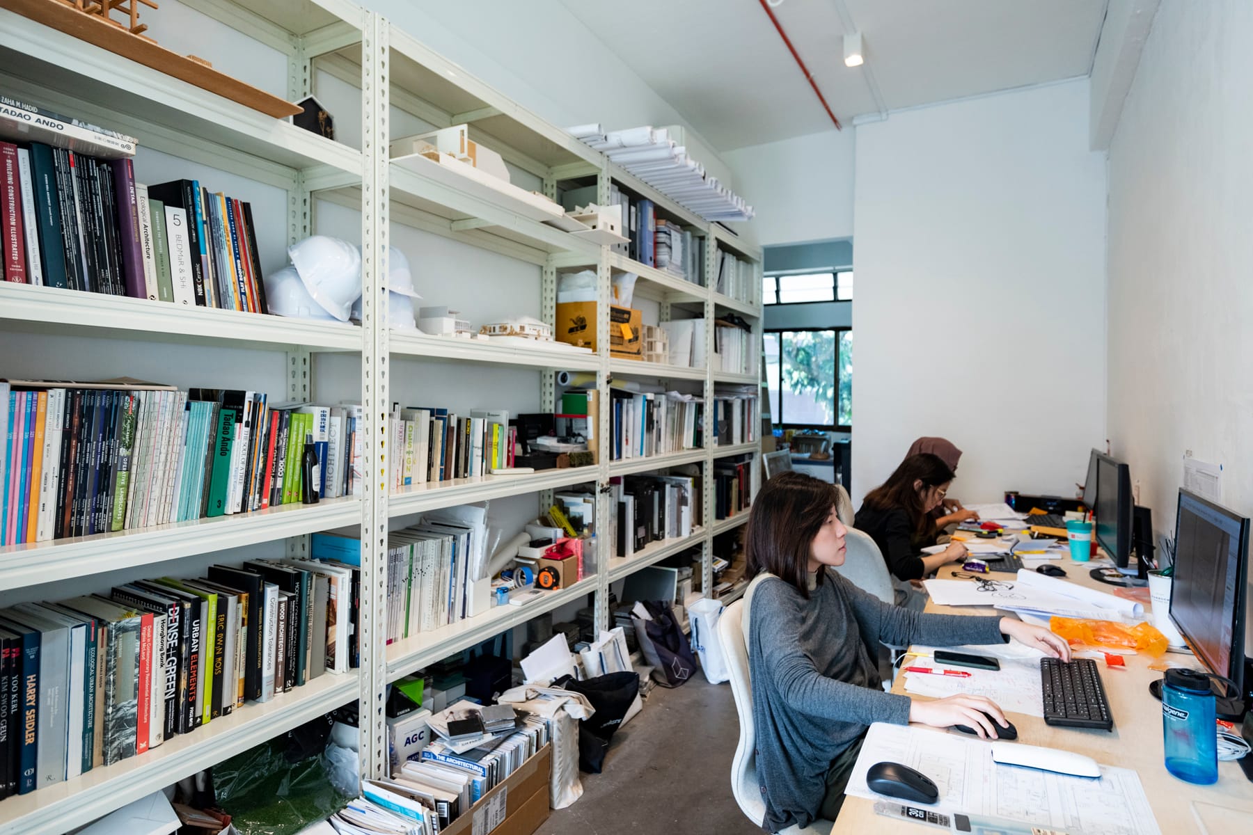Two people work at desks with computers in an office filled with tall bookshelves and various office items.