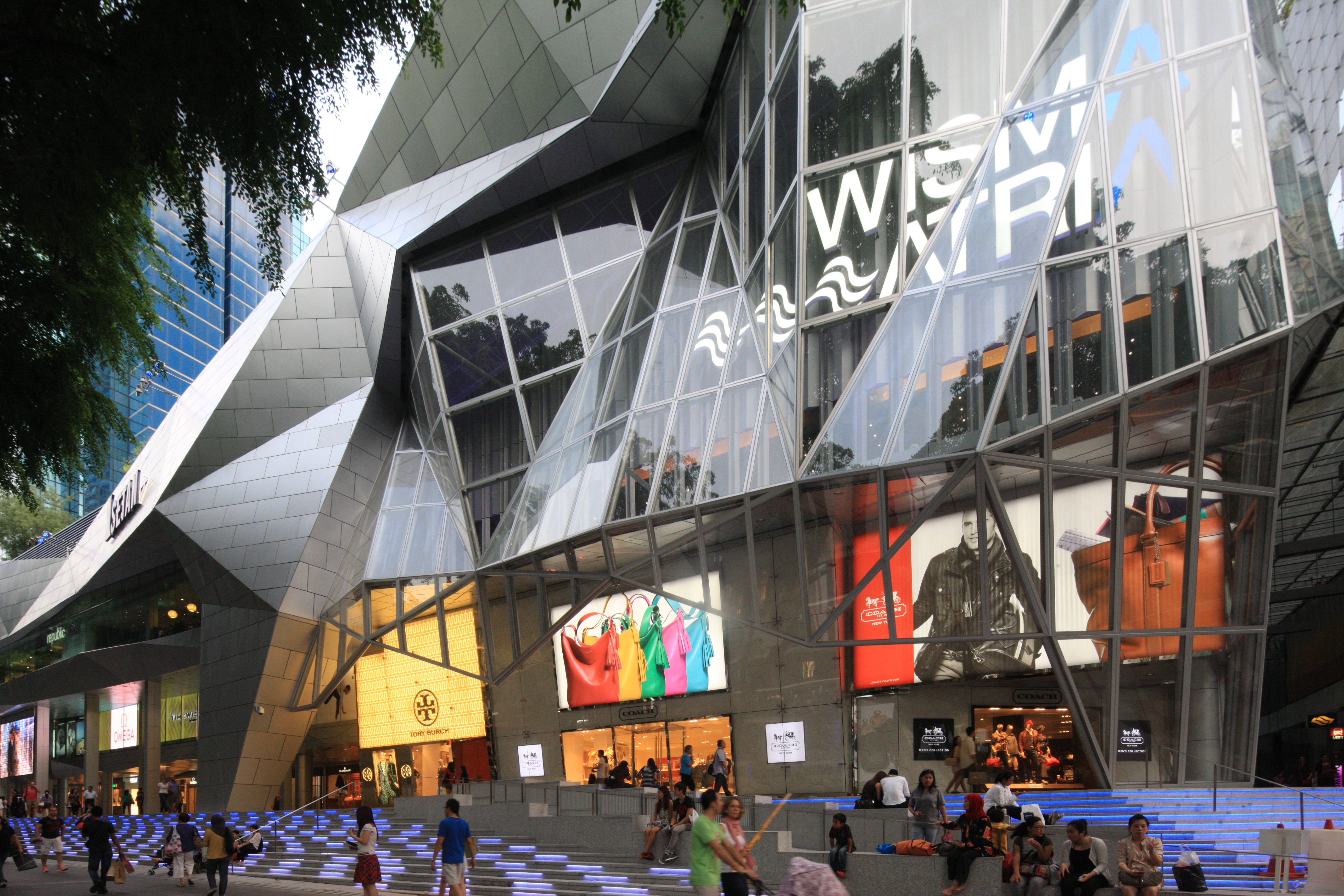 Modern glass building with angular design, storefronts displaying colorful bags and clothes, steps with blue lights, people walking.