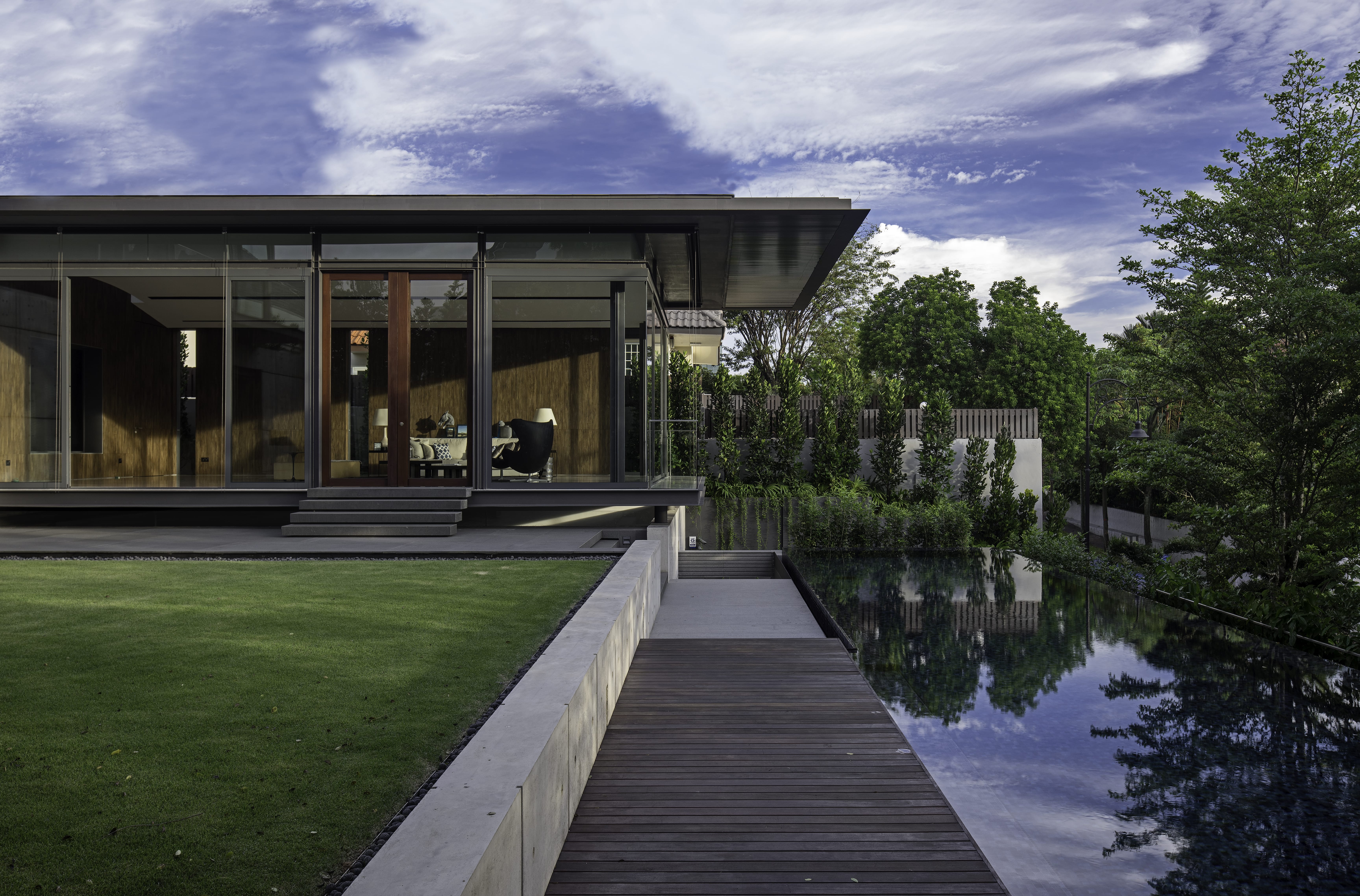 Modern glass-walled house with green lawn, wooden deck, and reflecting pool surrounded by trees under a partly cloudy sky.