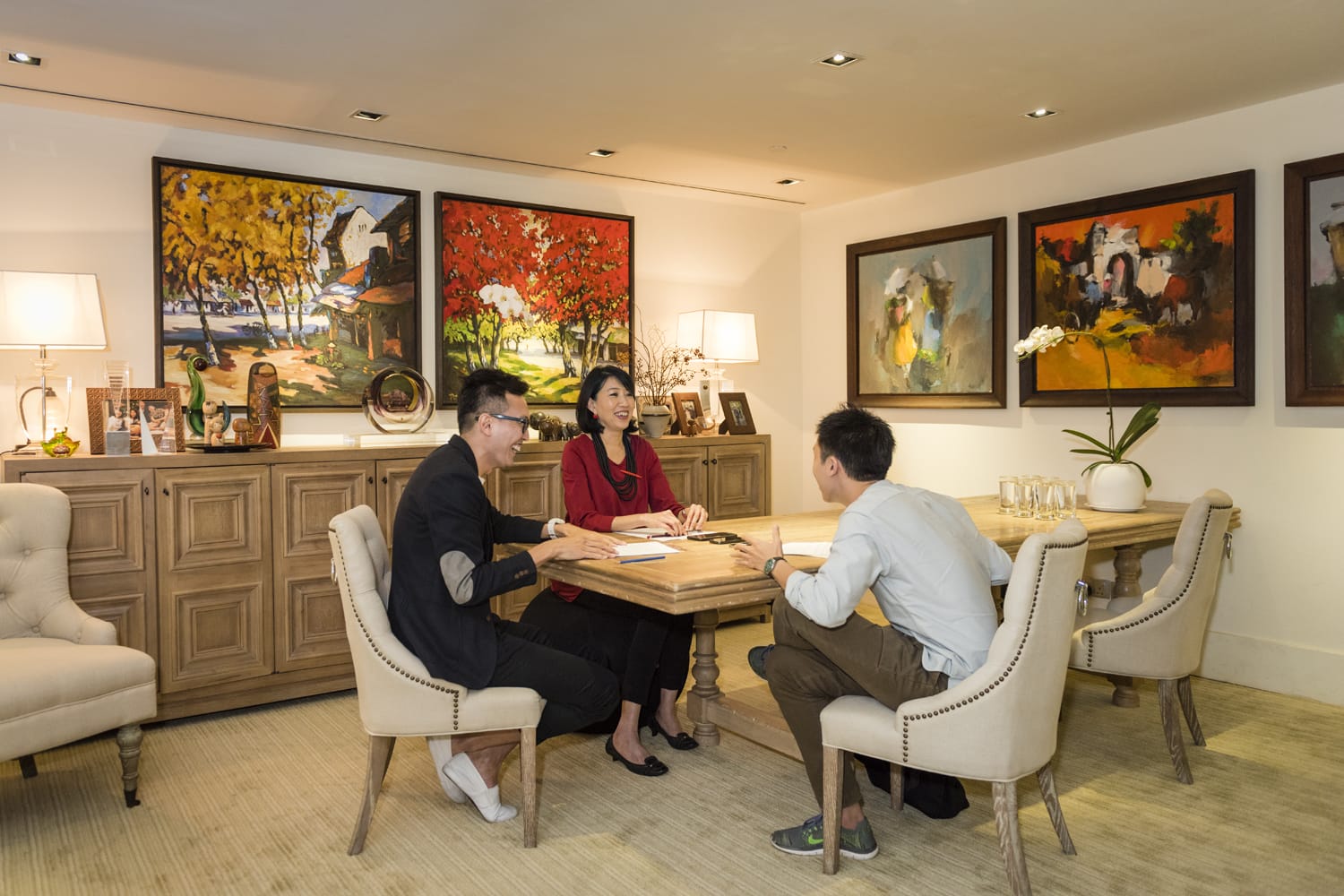 Three people sit at a wooden table in an art-adorned room with beige chairs and a sideboard.