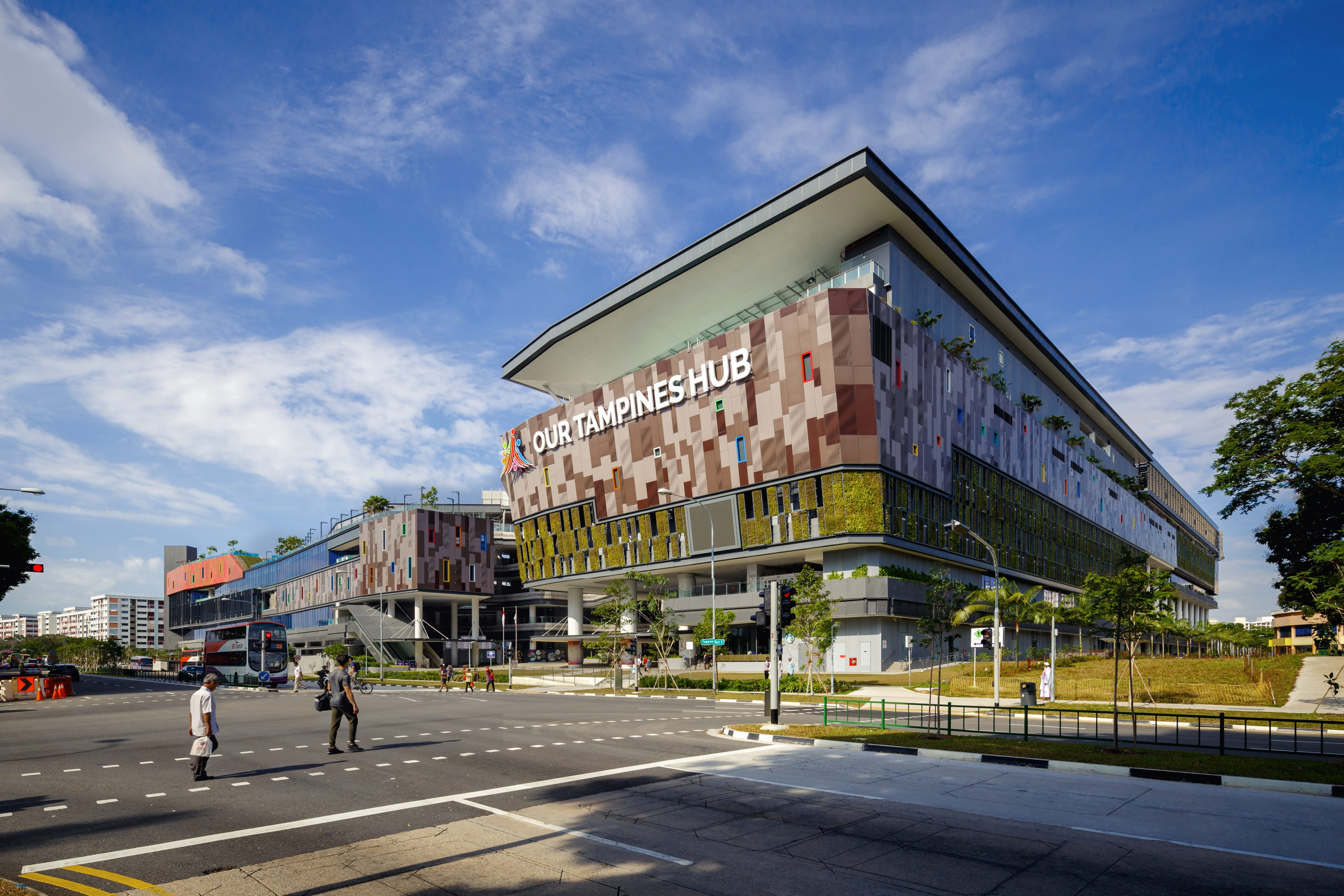 Modern building labeled "Our Tampines Hub" with crosswalk and pedestrians in the foreground on a sunny day.