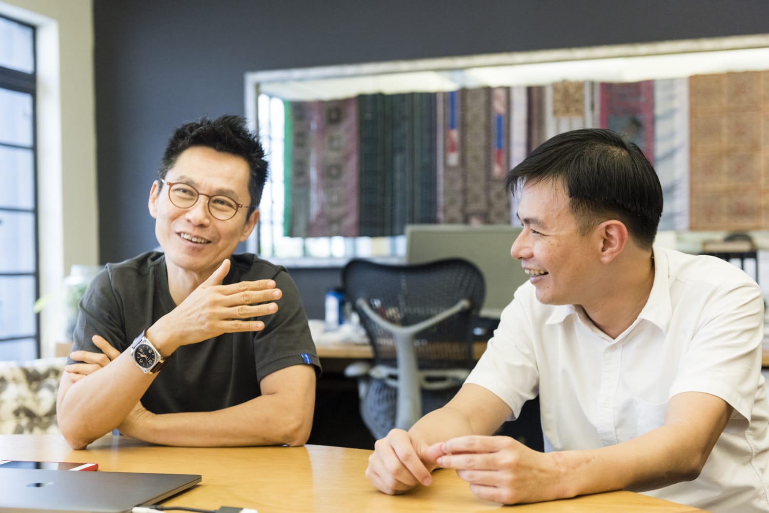 Two people seated at an office table, one gesturing with their hand, with a laptop and documents in front of them.