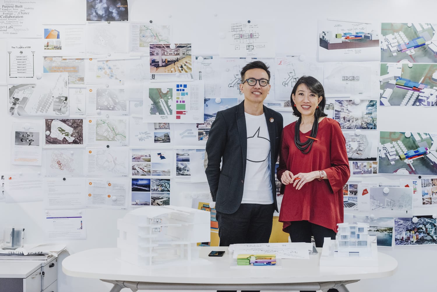 Two people stand in front of a wall covered with architectural plans and models on a desk.