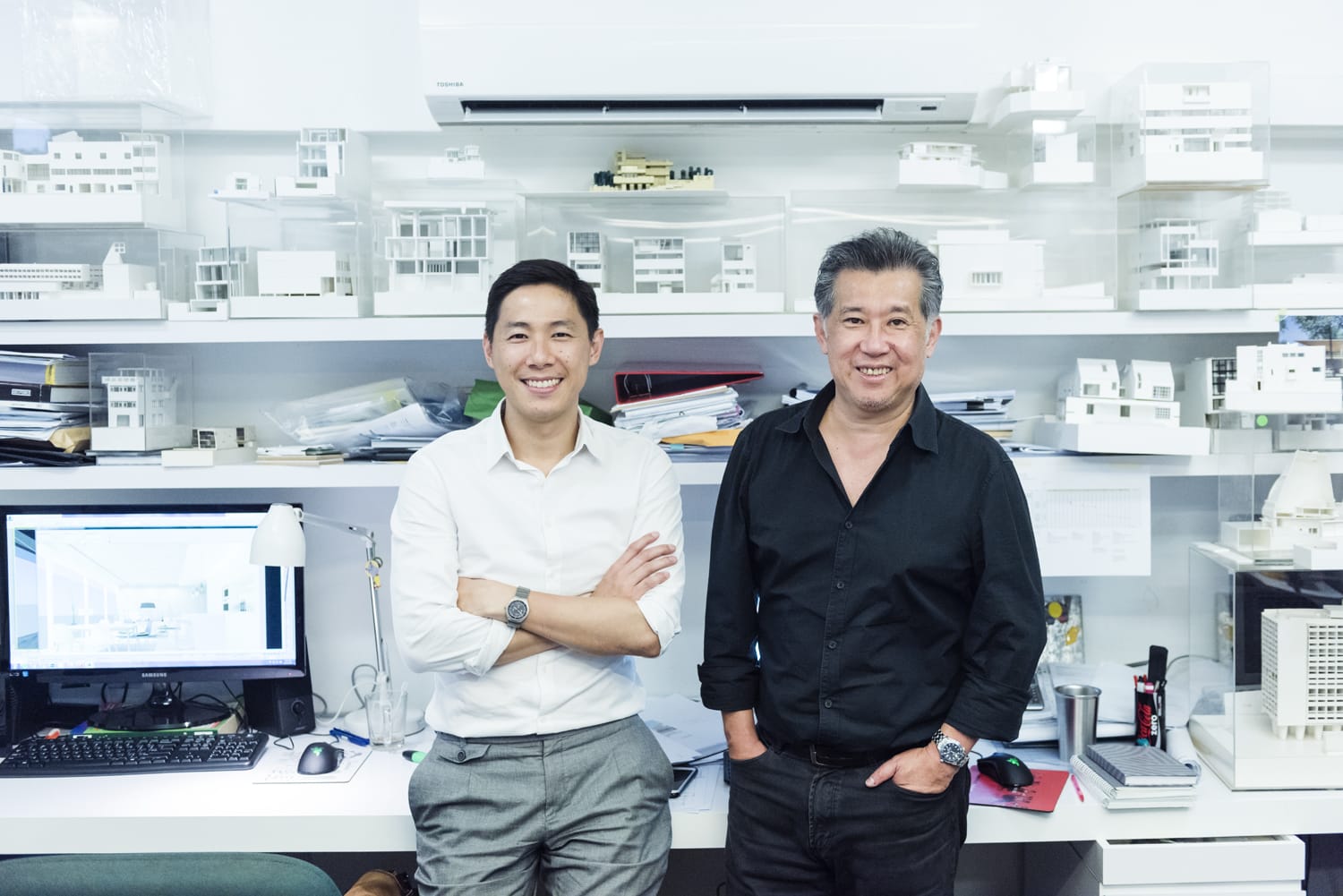 Two people stand in an office with white shelves displaying architectural models and a desk with a computer.