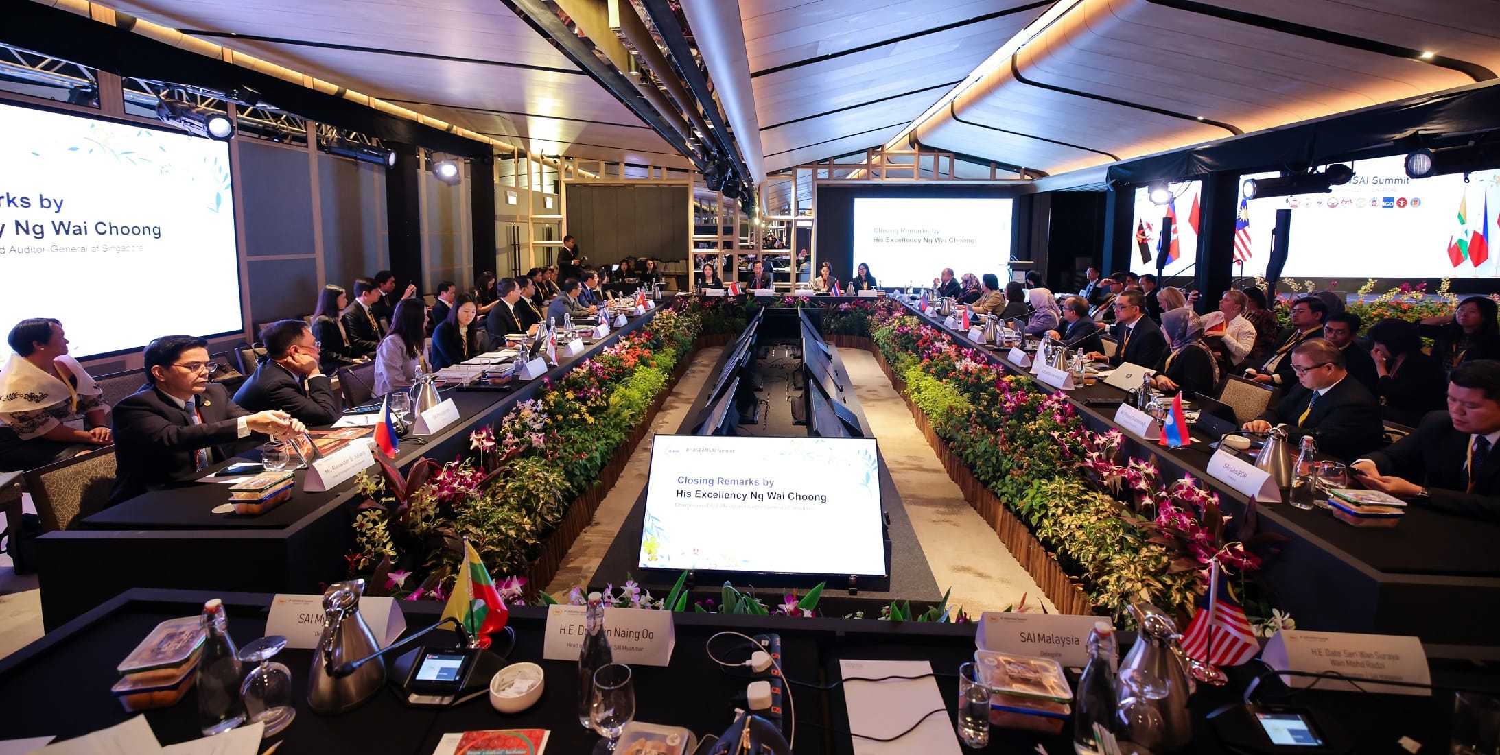 Large conference room with participants seated around a U-shaped table, decorated with flags and flower arrangements.