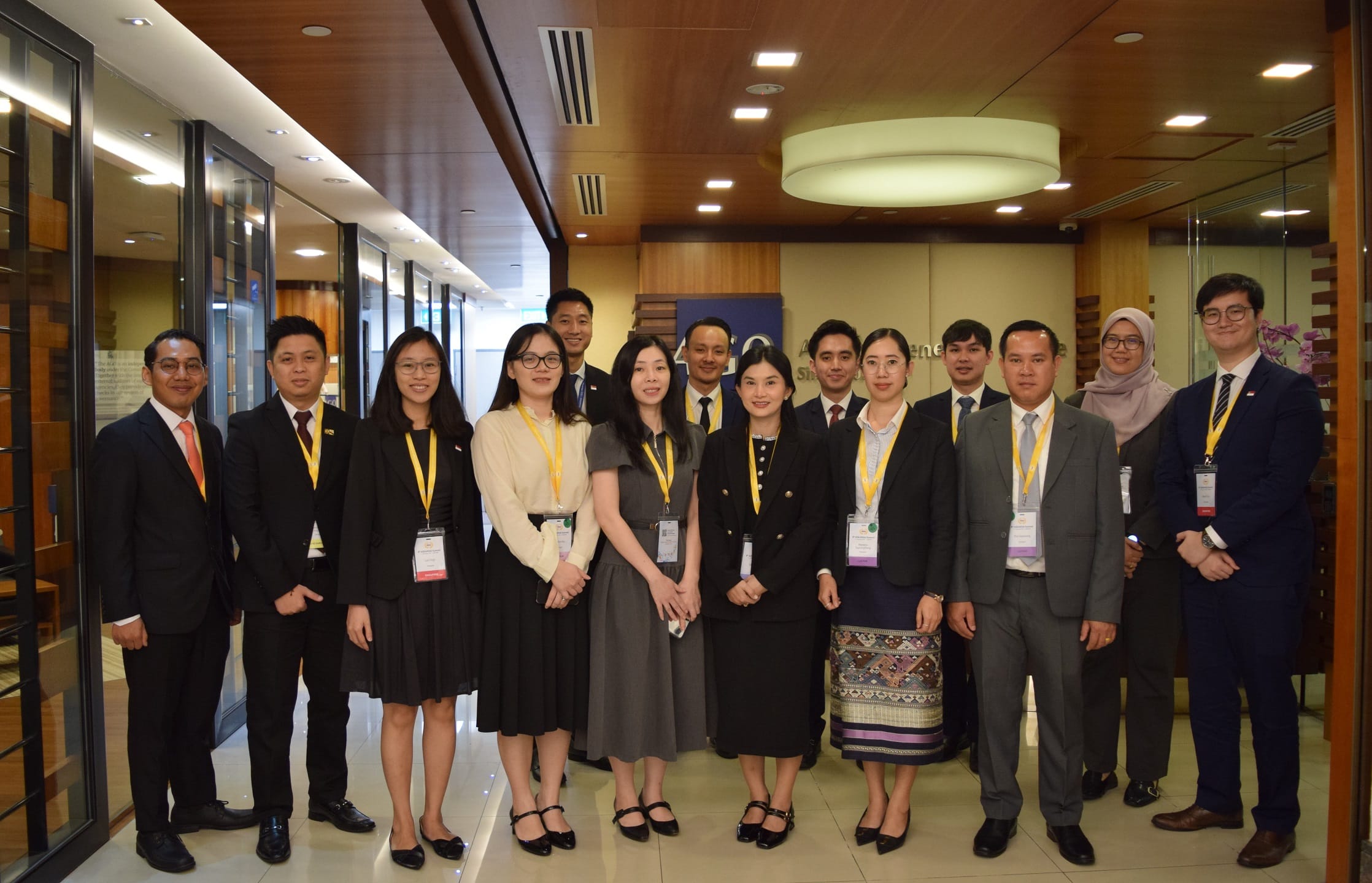 A group of people in formal attire and name tags posing in a hallway with wooden ceiling and glass walls.