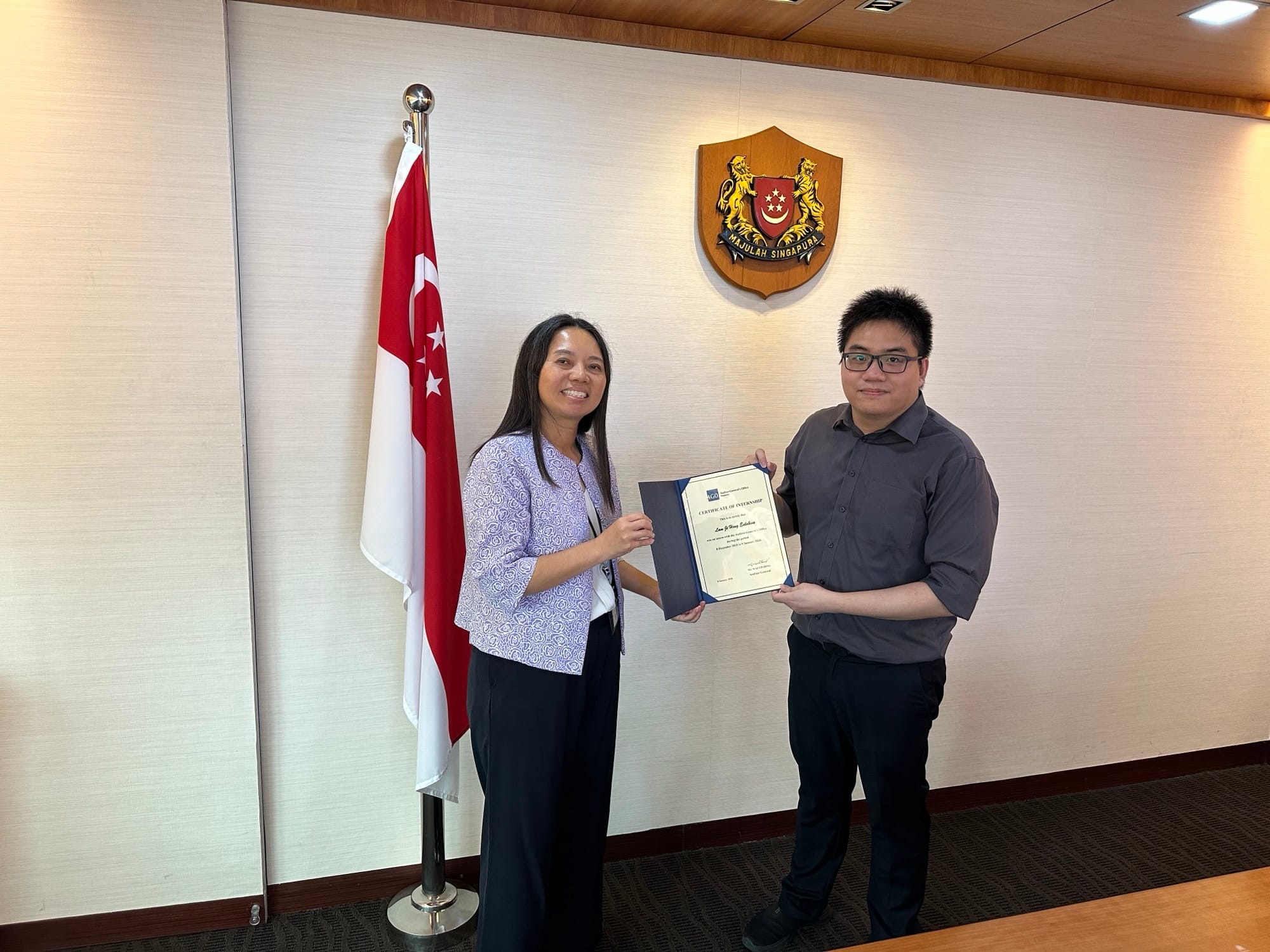 Two people standing under the Singapore crest, holding a certificate, next to the Singapore flag.