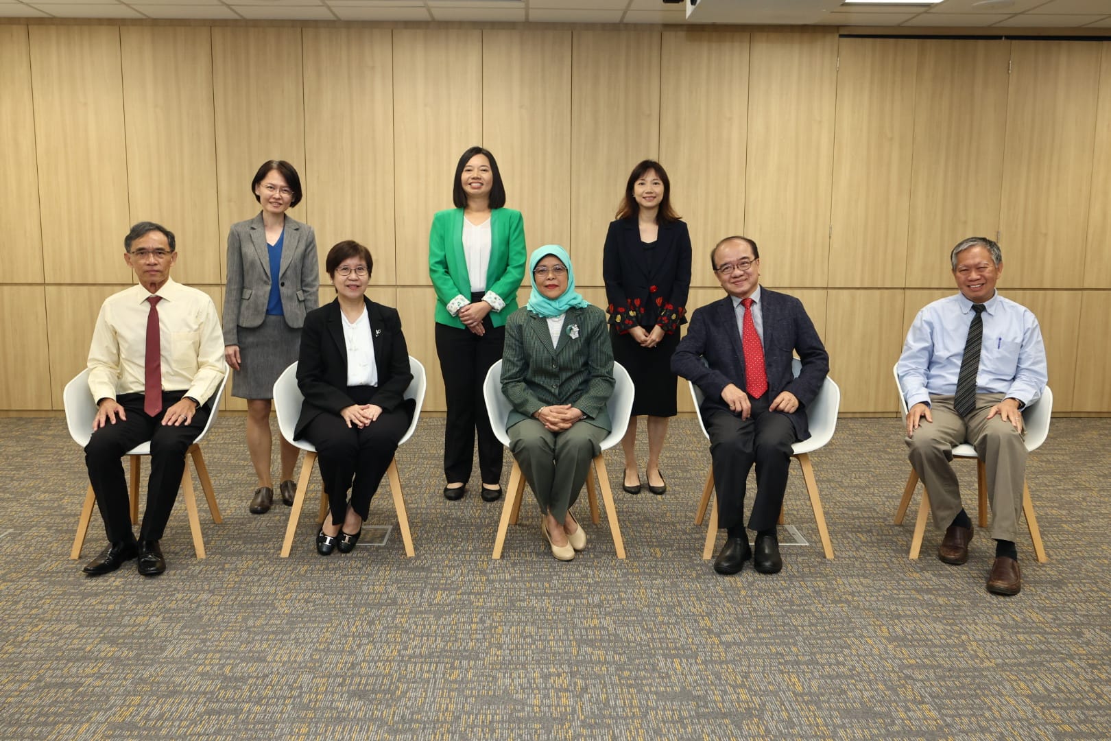 Eight people in business attire are posing in front of a wooden wall, with three standing and four seated.