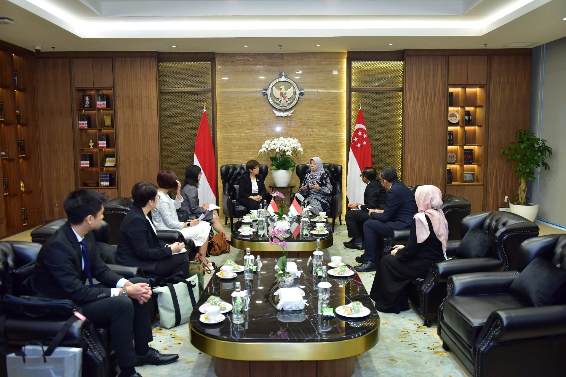 People seated in a formal meeting room with Indonesian and Singaporean flags, a large table, and bookshelf visible.