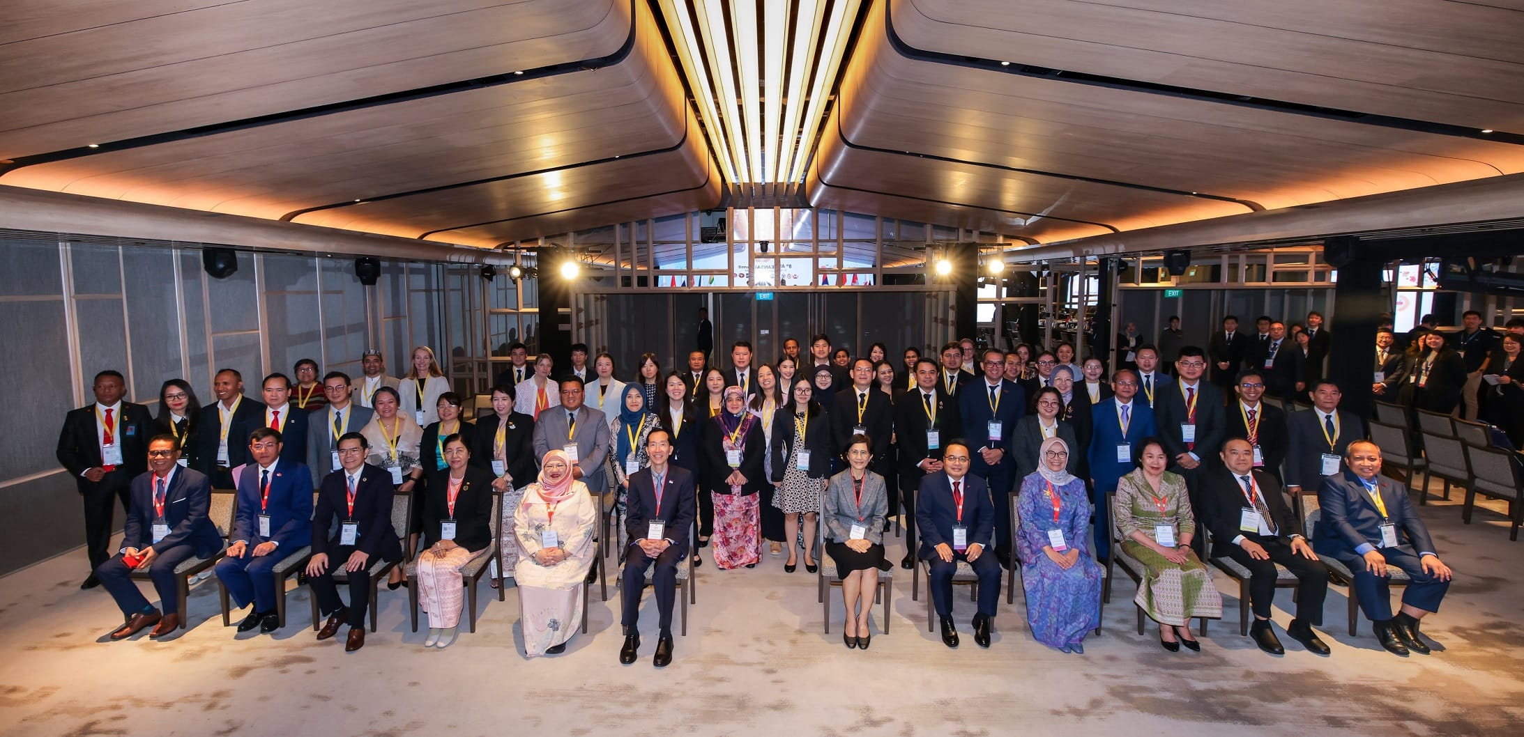 A large group of people in formal attire are seated and standing in a modern conference room.