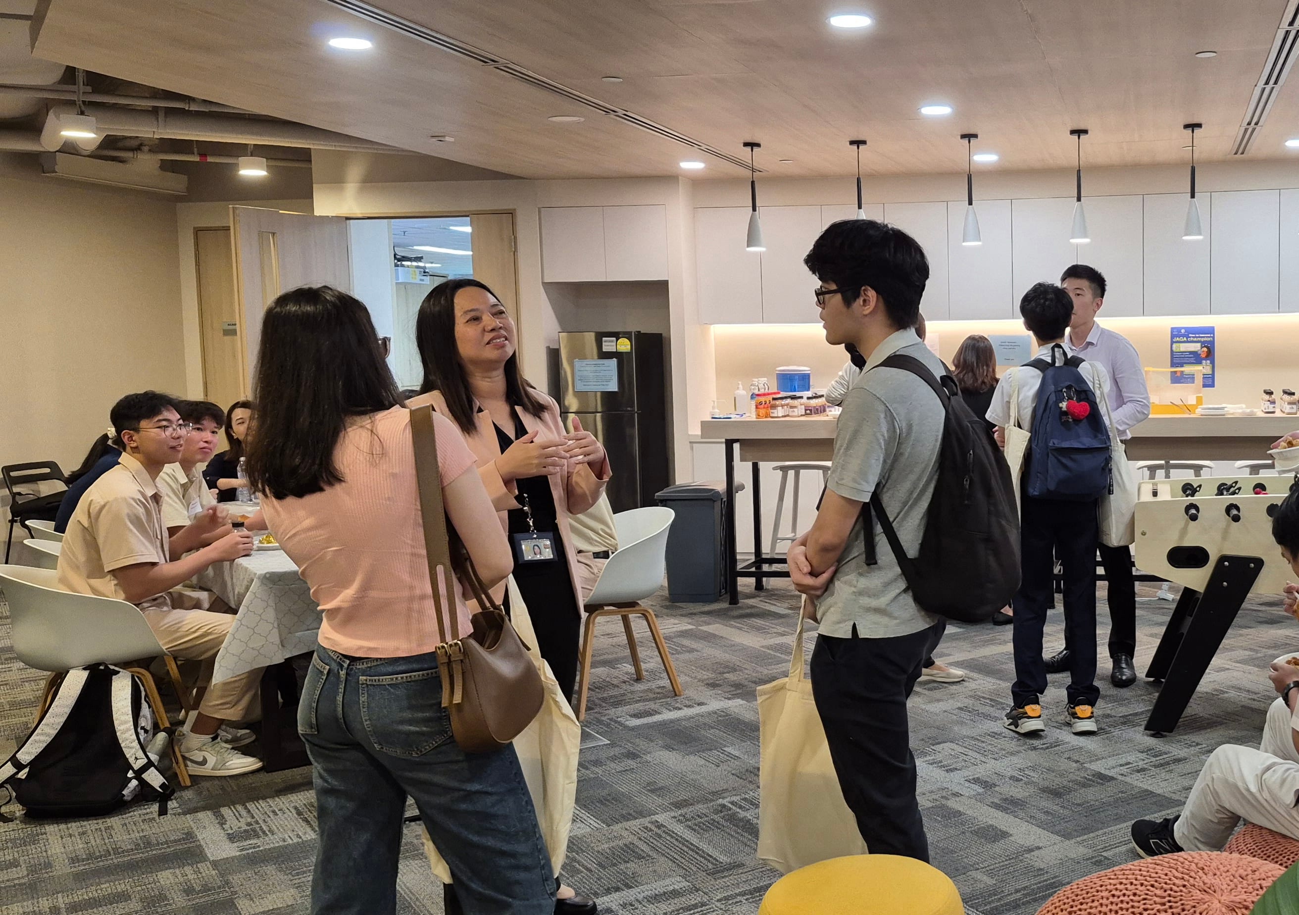 People are gathered and interacting in a modern office area with tables, chairs, and a foosball table in the background.