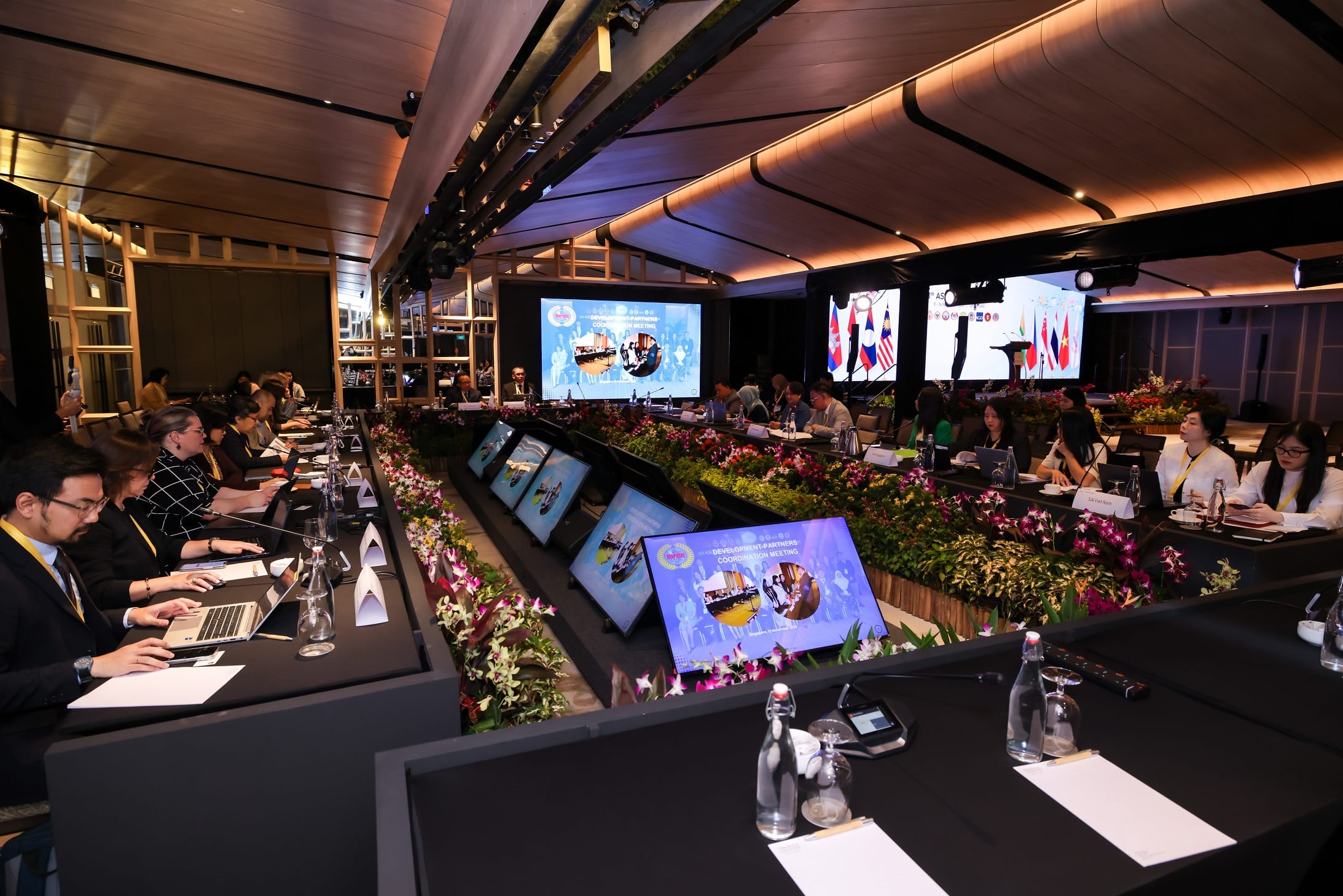 Conference room with attendees seated at long tables with laptops, large screens displaying images, and decorated with flowers.