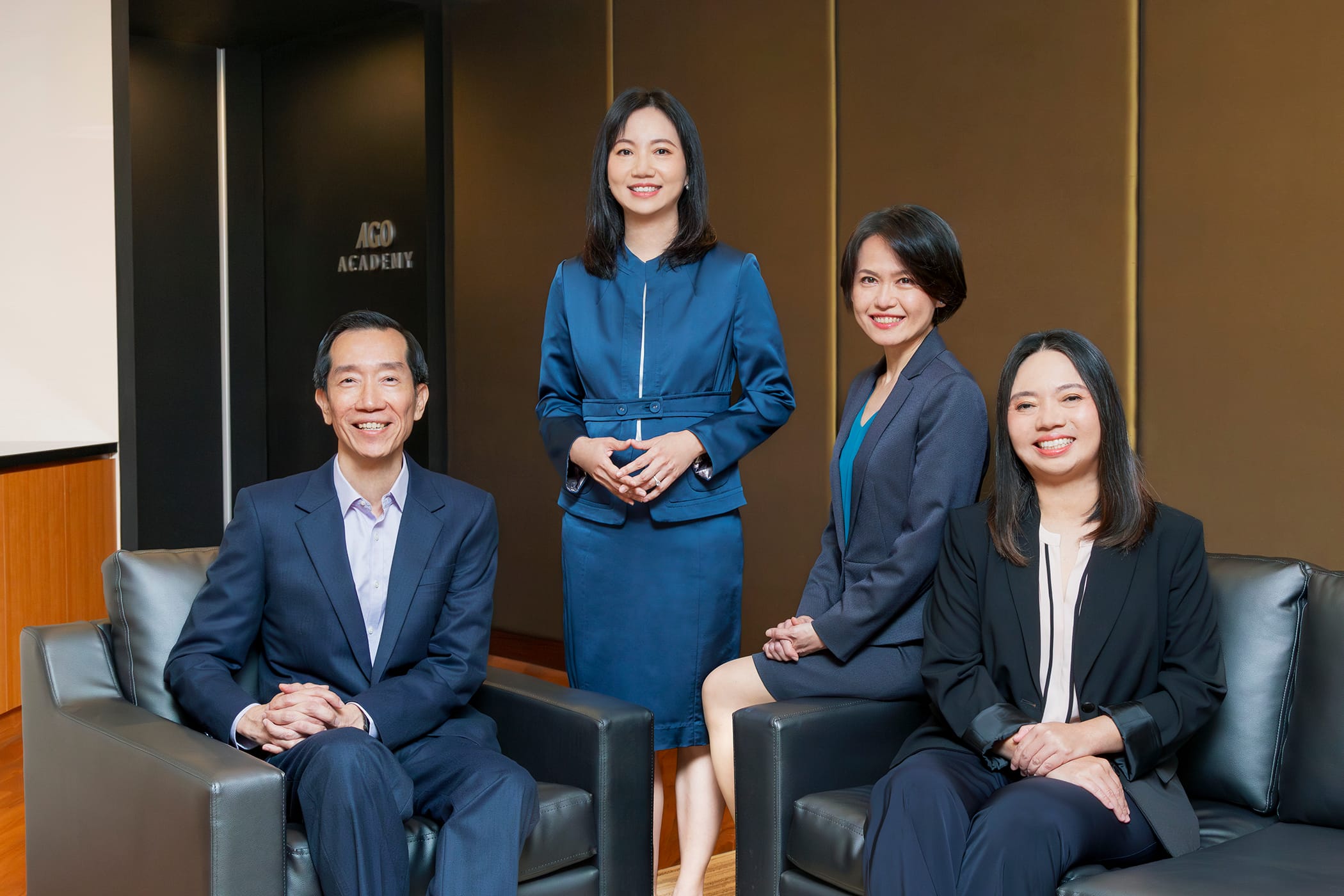 Four people in formal business attire are posing in an office setting with "AGO Academy" written on the wall.