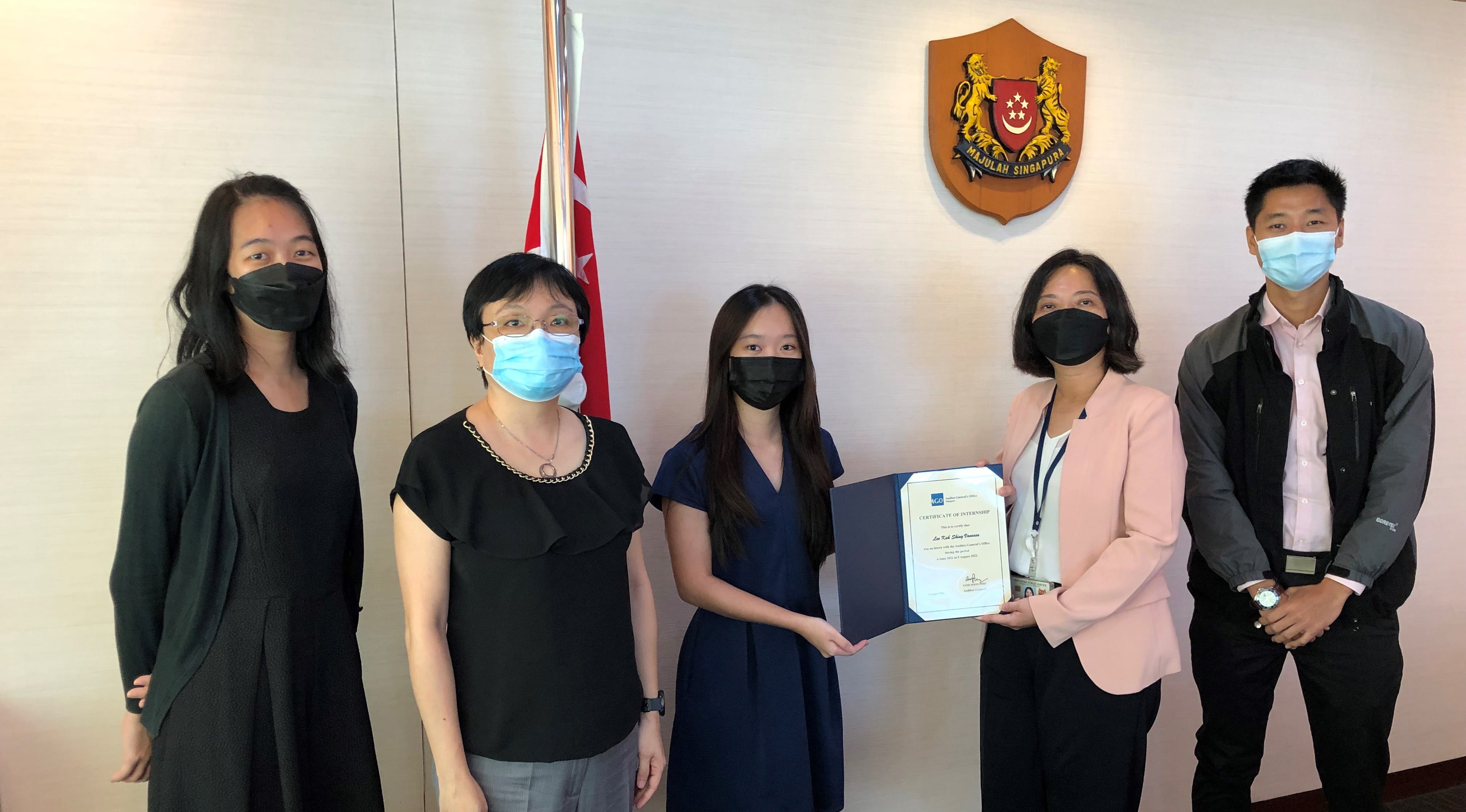 Five individuals are standing in front of a wall with the Singapore crest and flag, and two people holding a certificate.