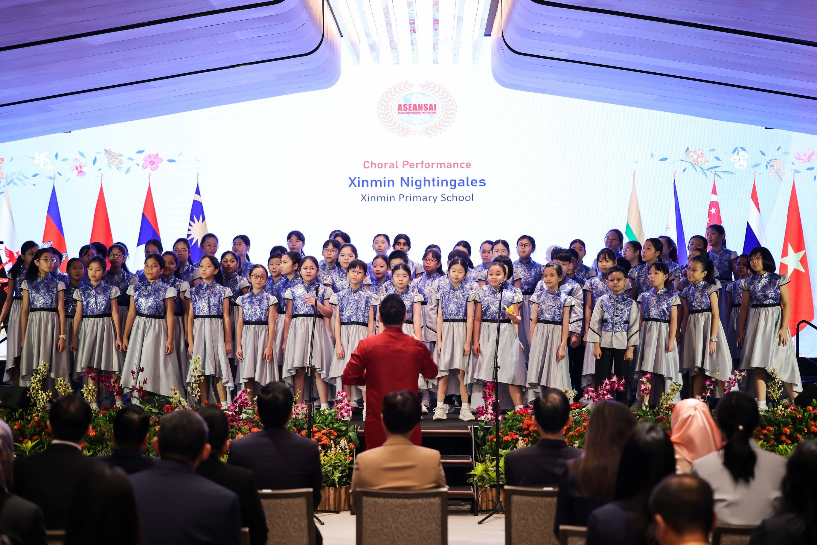 A large group of children performs on stage wearing coordinated blue and gray outfits, directed by a person in red, facing an audience.