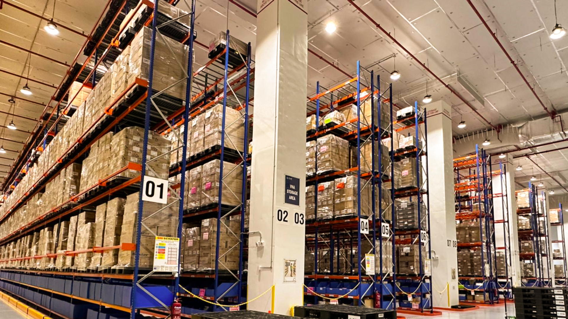 Warehouse with tall shelves holding stacked pallets of boxes, numbered sections, and a sign reading "VNA STORAGE AREA."