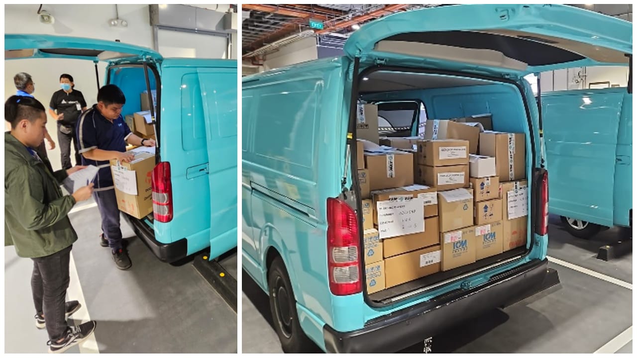 A turquoise van with its rear doors open, loaded with numerous cardboard boxes, and two people examining paperwork.