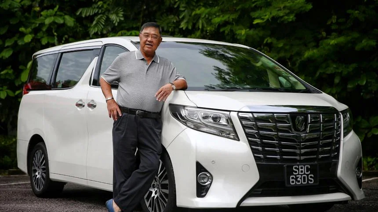 A person is wearing a gray polo shirt, dark pants, and slippers, standing next to a white minivan with license plate SBC 630S.