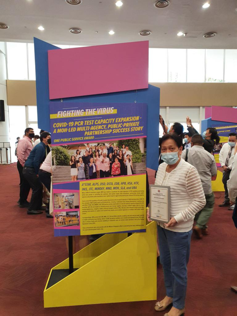 Person holding a certificate stands beside a large display board about COVID-19 PCR test capacity expansion and partnerships.