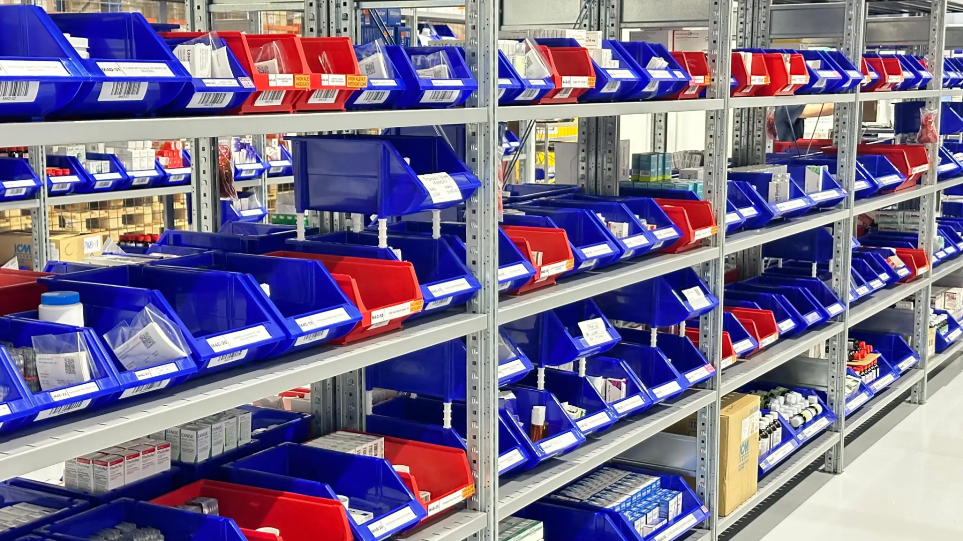 Shelving unit with blue and red plastic bins containing various medical supplies in a warehouse setting.