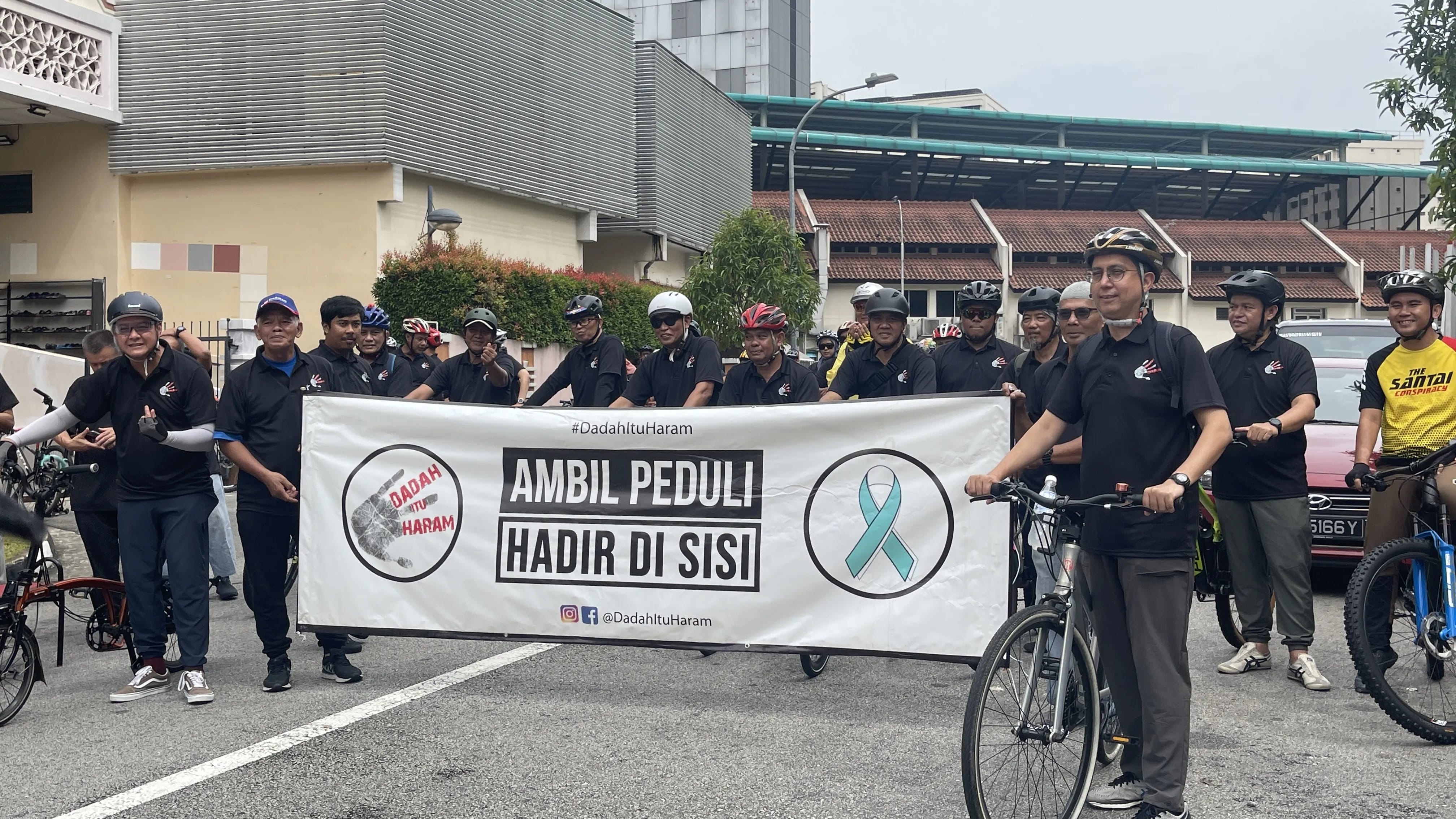 A group of cyclists in black shirts and helmets holding a banner with anti-drug messages in Indonesian.